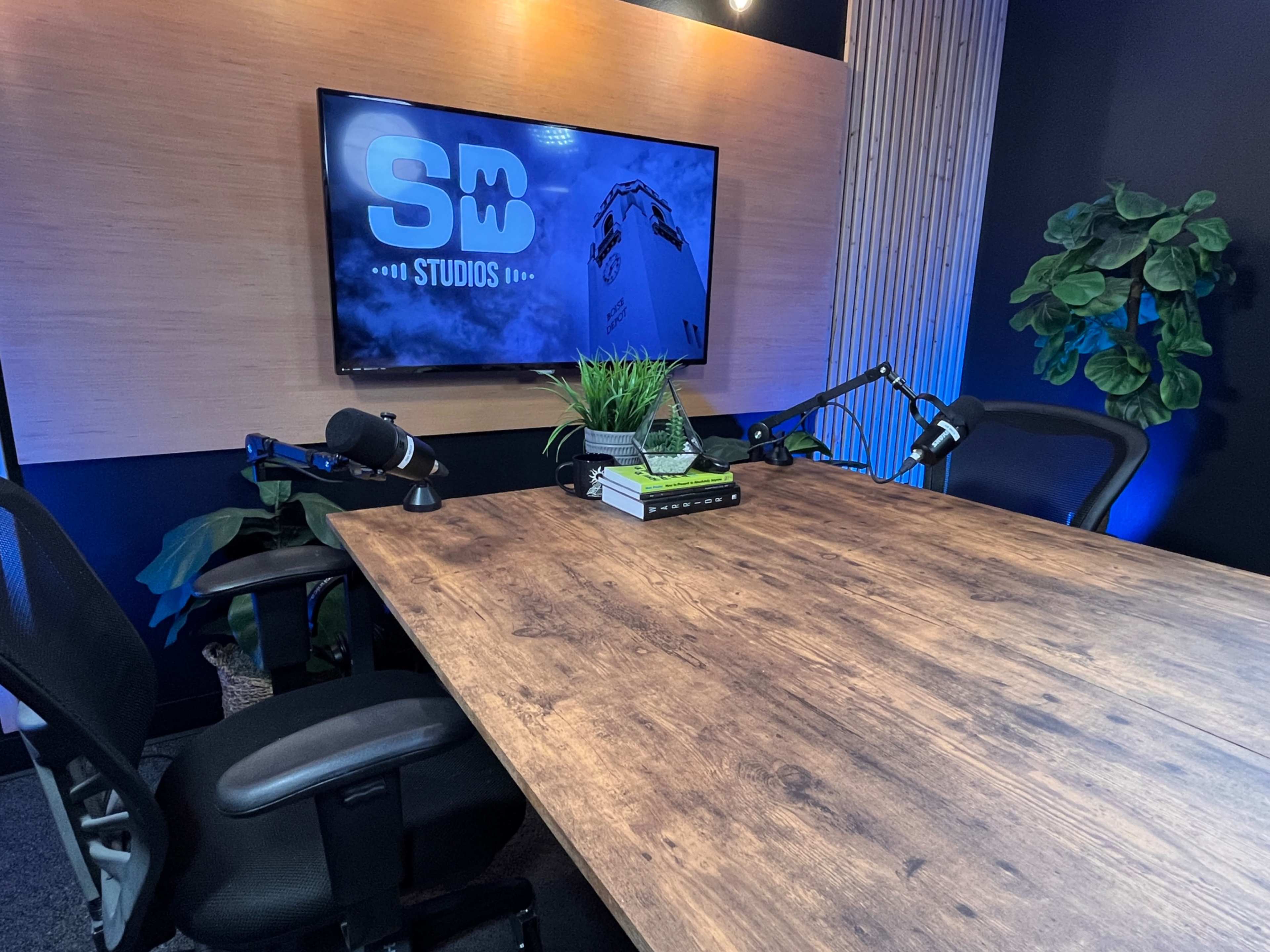 The image shows a modern studio setup with a wooden table, two black office chairs, and a large screen displaying "SW Studios," alongside plants and microphones on the table.