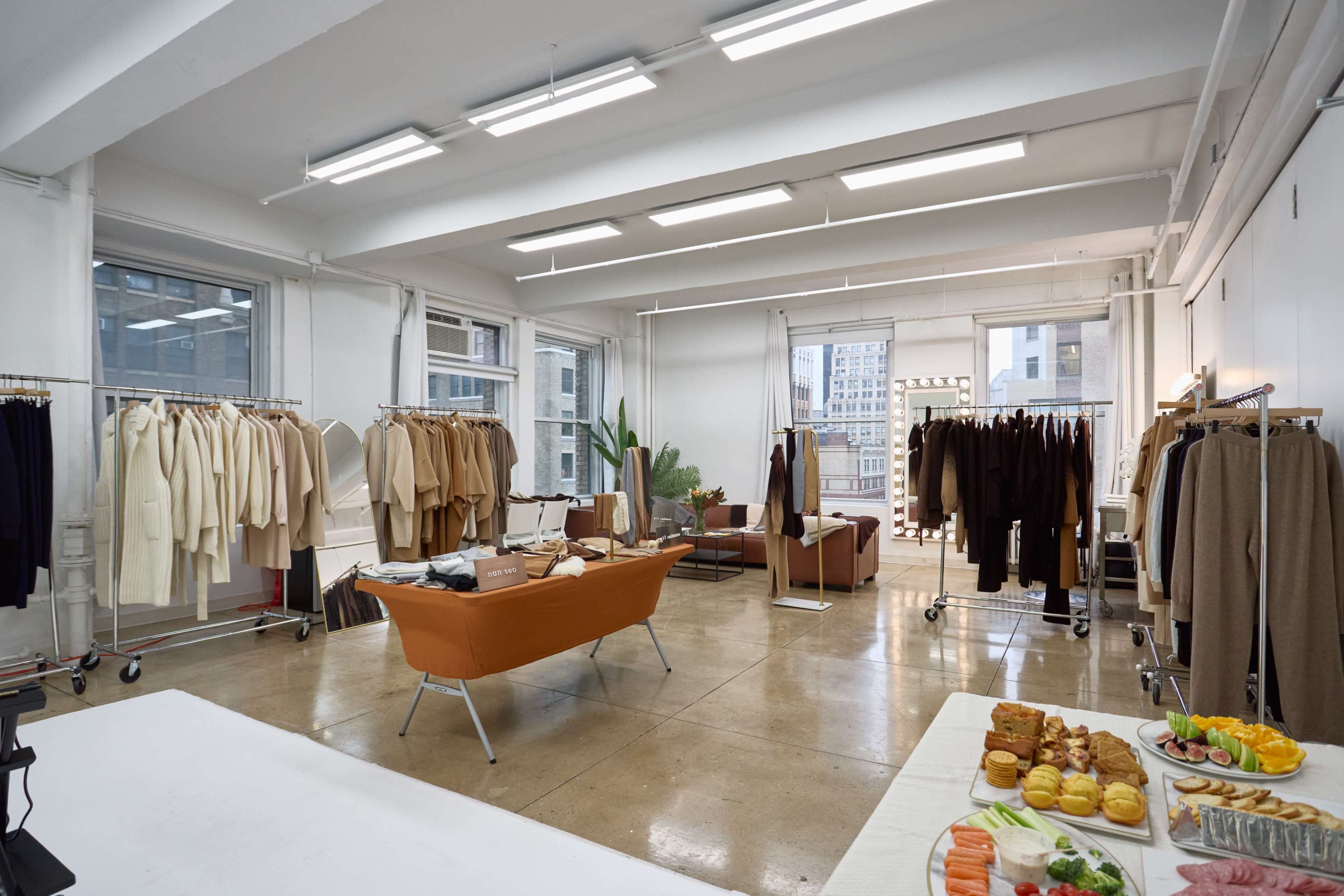 A spacious studio displays racks of clothing in neutral colors, with a seating area and a table of assorted food in the foreground.