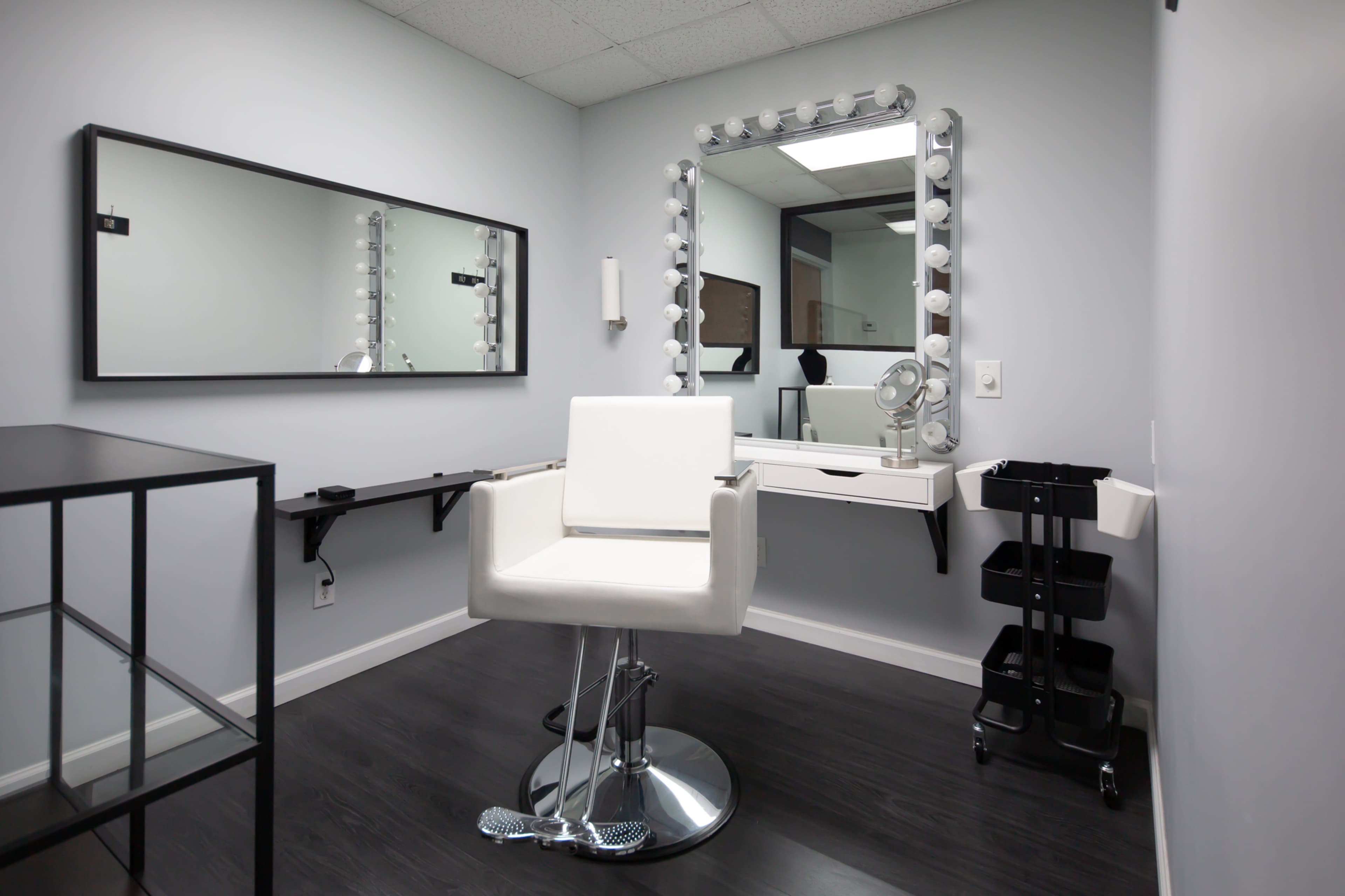 A modern beauty salon room featuring a makeup chair, illuminated mirror, and minimalist decor.