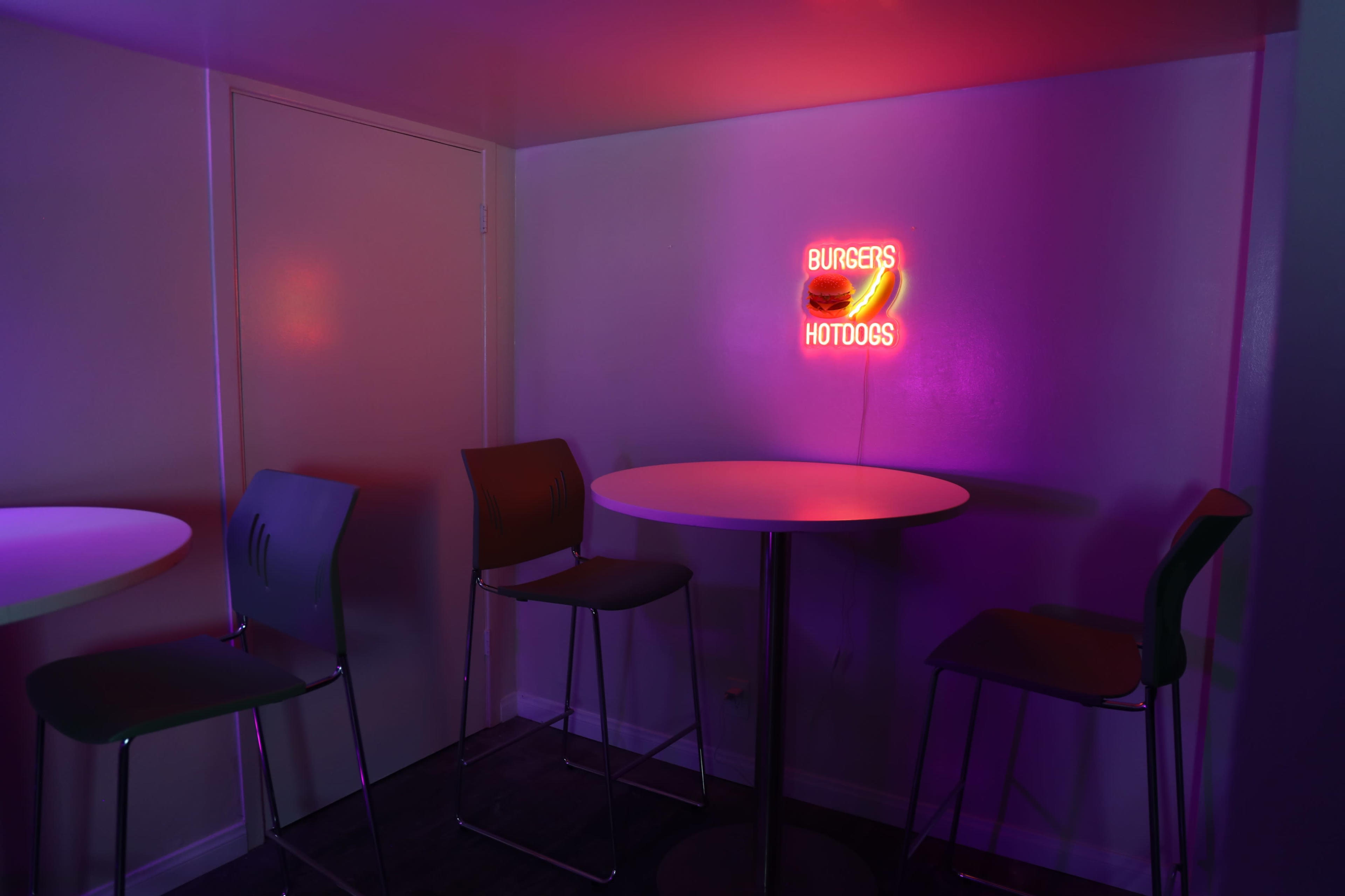 A small seating area features a round table and three metal chairs under a neon sign that reads "BURGERS HOTDOGS."