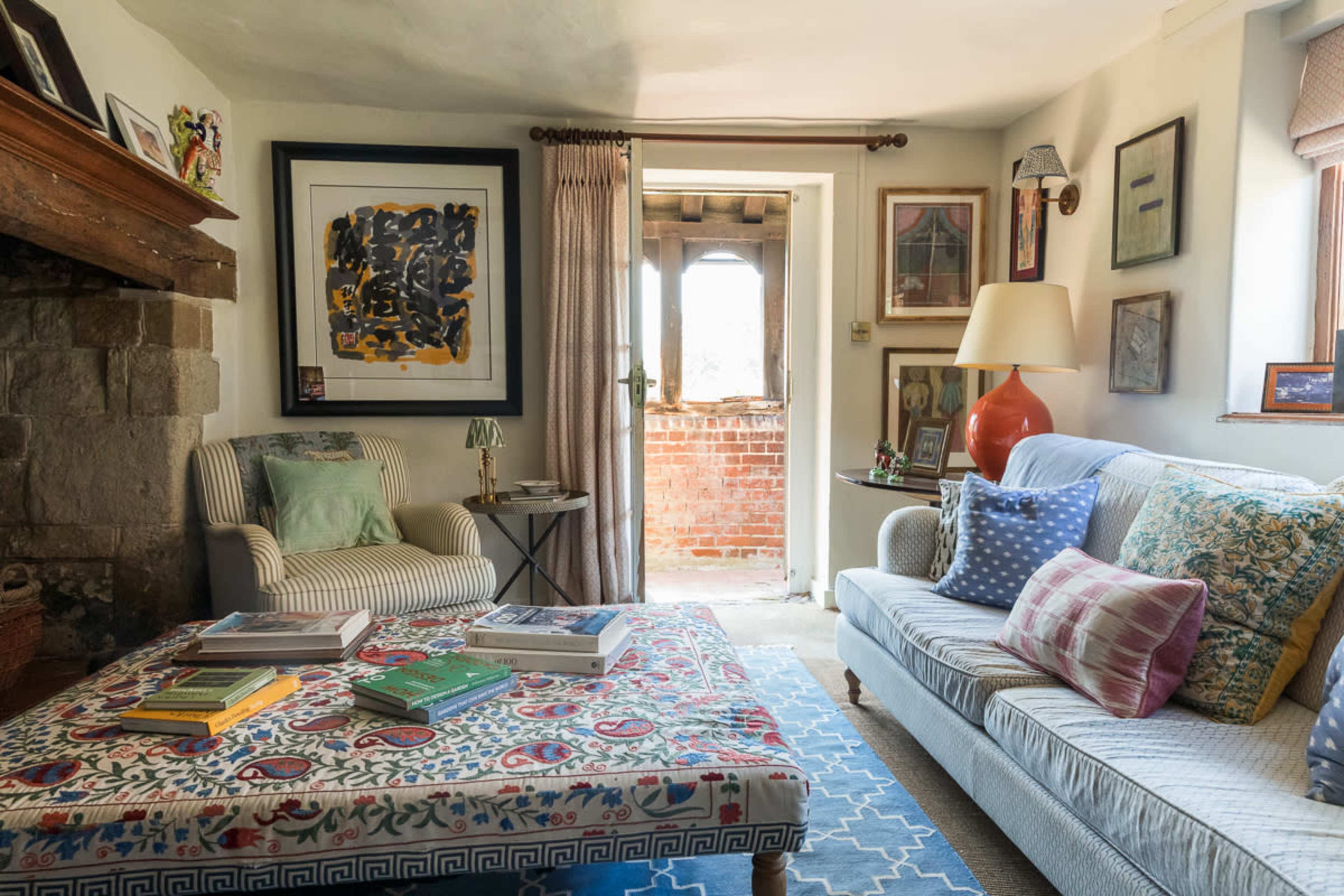 The image shows a cozy living room featuring a patterned sofa, a striped armchair, a coffee table with books, and a doorway leading to a brick balcony.