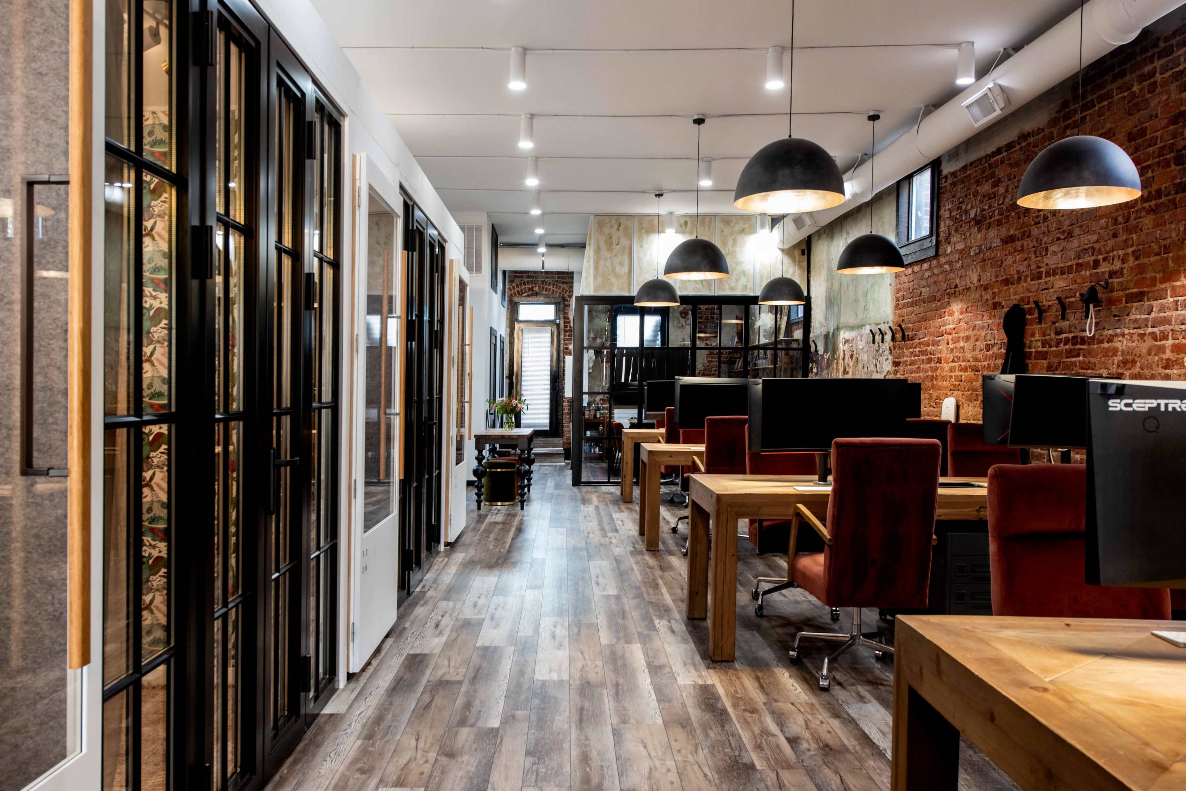 A modern open office space with wooden desks, black chairs, and glass partitioned meeting rooms along a corridor with exposed brick walls.