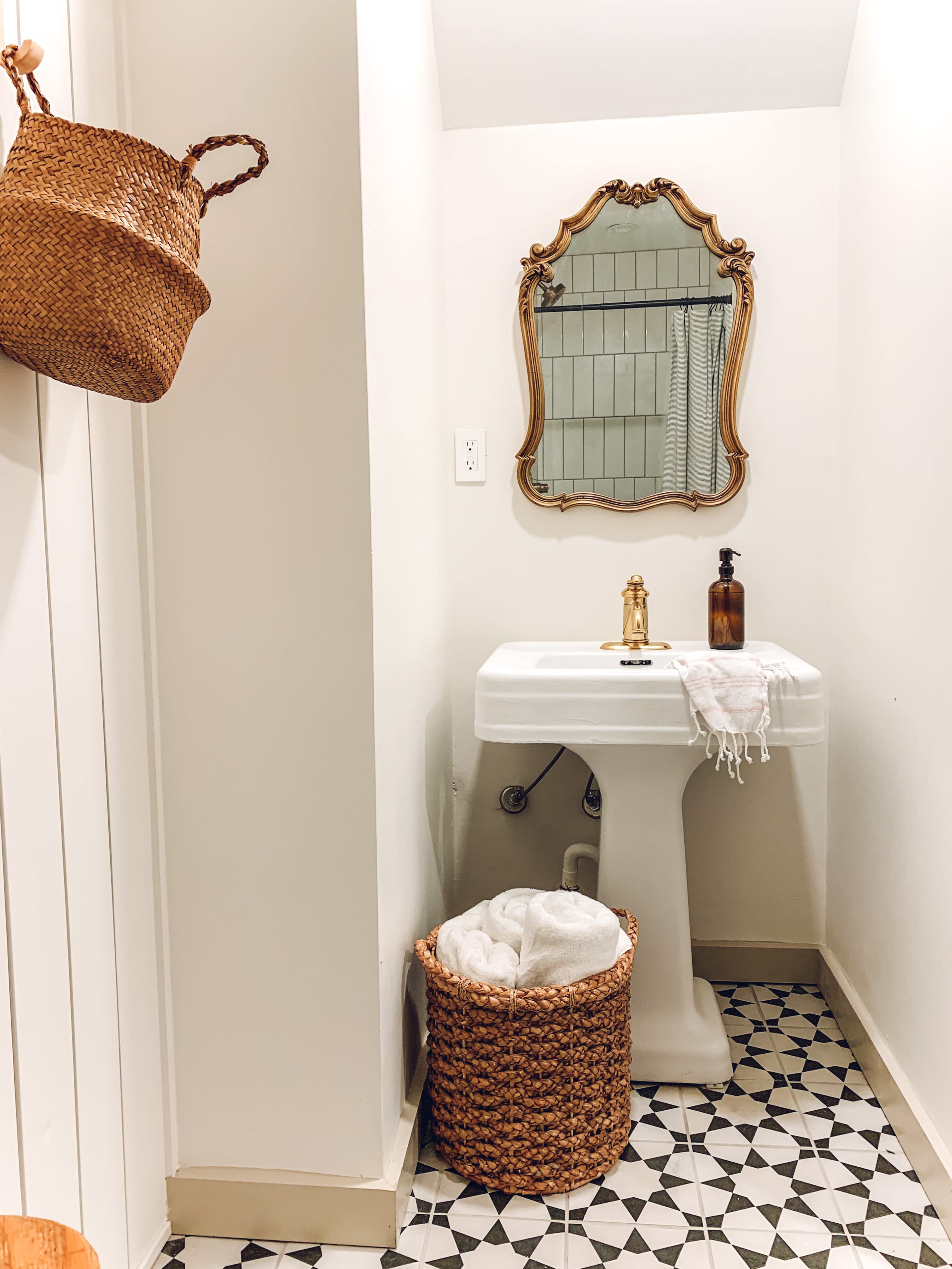 A small bathroom features a vintage-style mirror above a pedestal sink, with woven baskets hanging on the wall and a patterned tile floor.