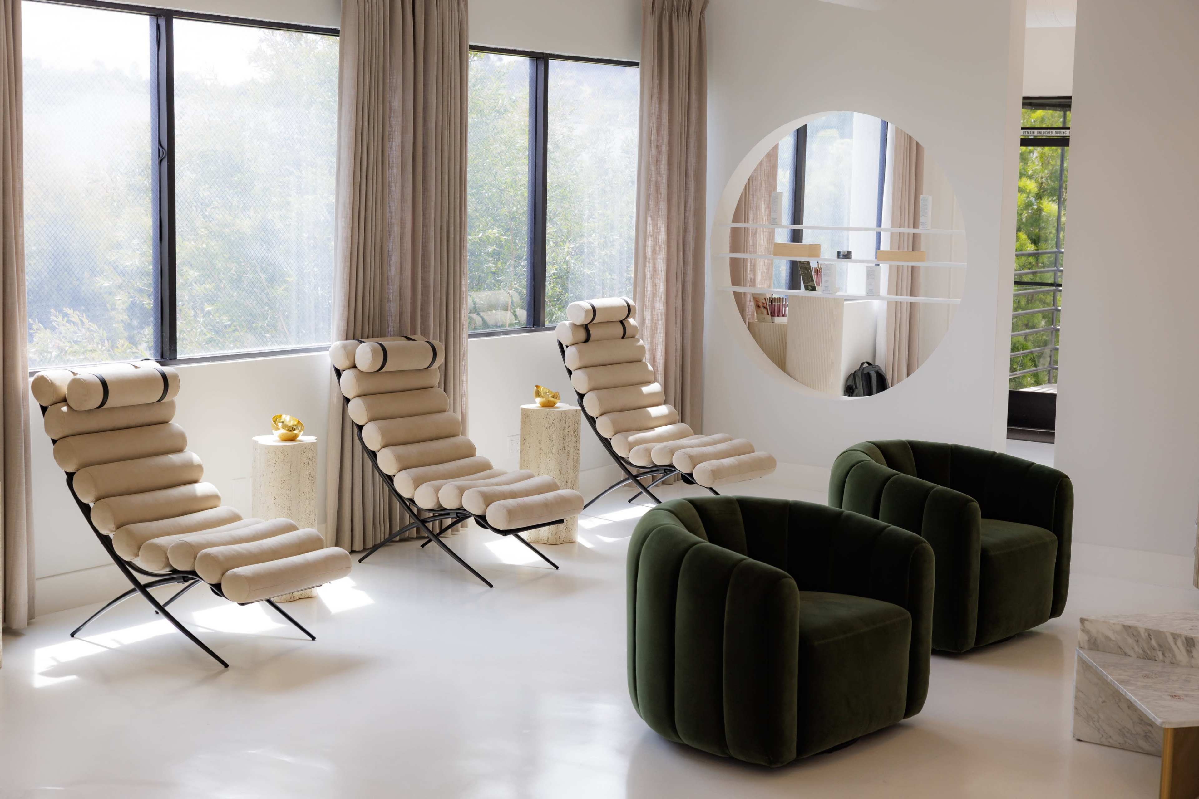 The image shows a modern living room with three beige lounge chairs and two green armchairs near large windows draped with light curtains.