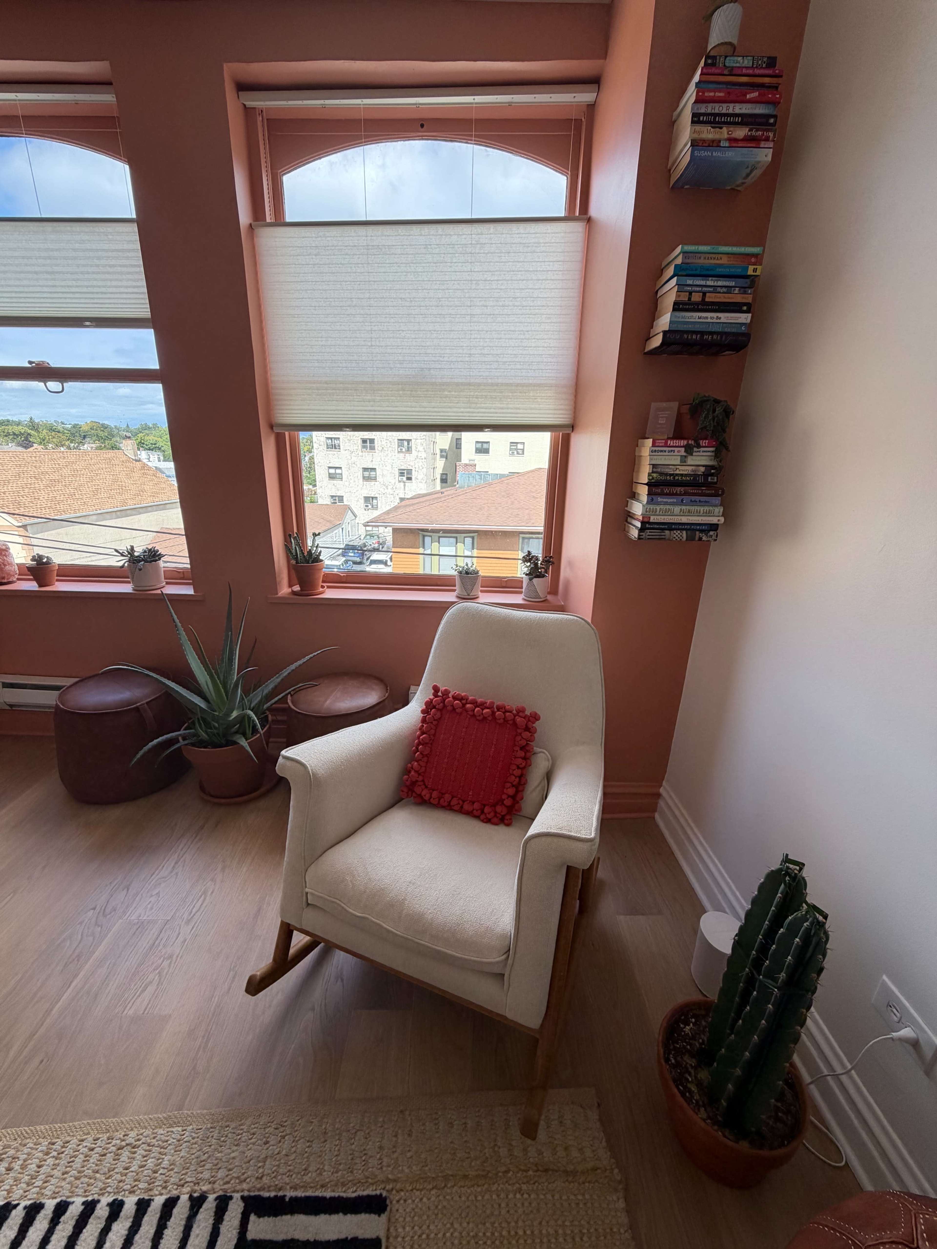 A cozy corner features a white armchair with a red cushion, surrounded by potted plants and a wall-mounted bookshelf filled with books.