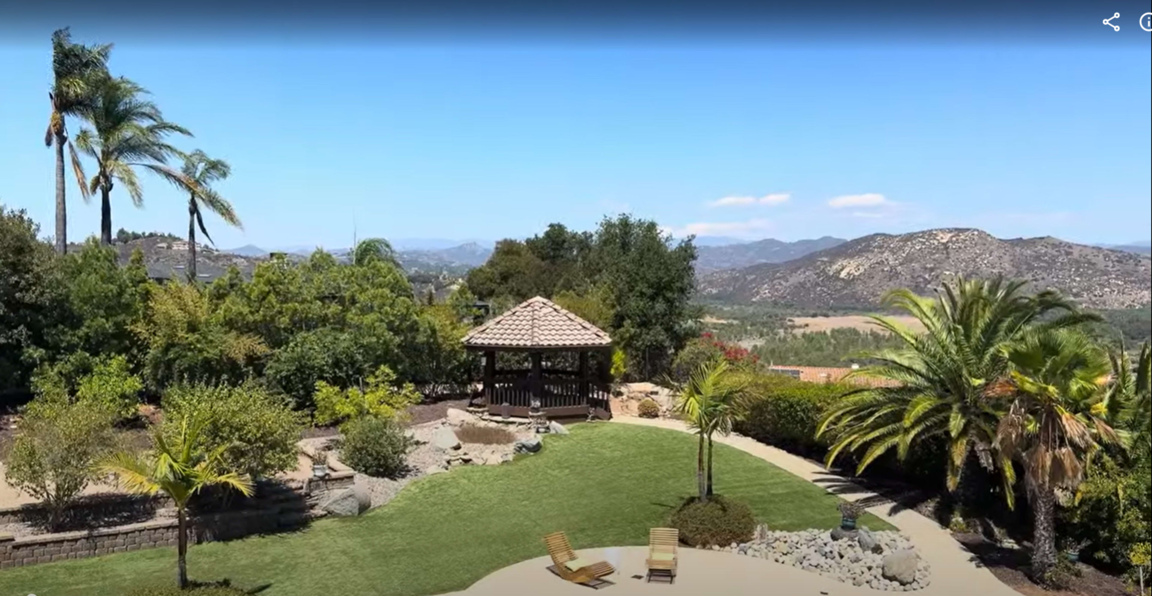 The image shows a landscaped yard with a gazebo and two lounge chairs, surrounded by trees and mountains in the background under a clear blue sky.