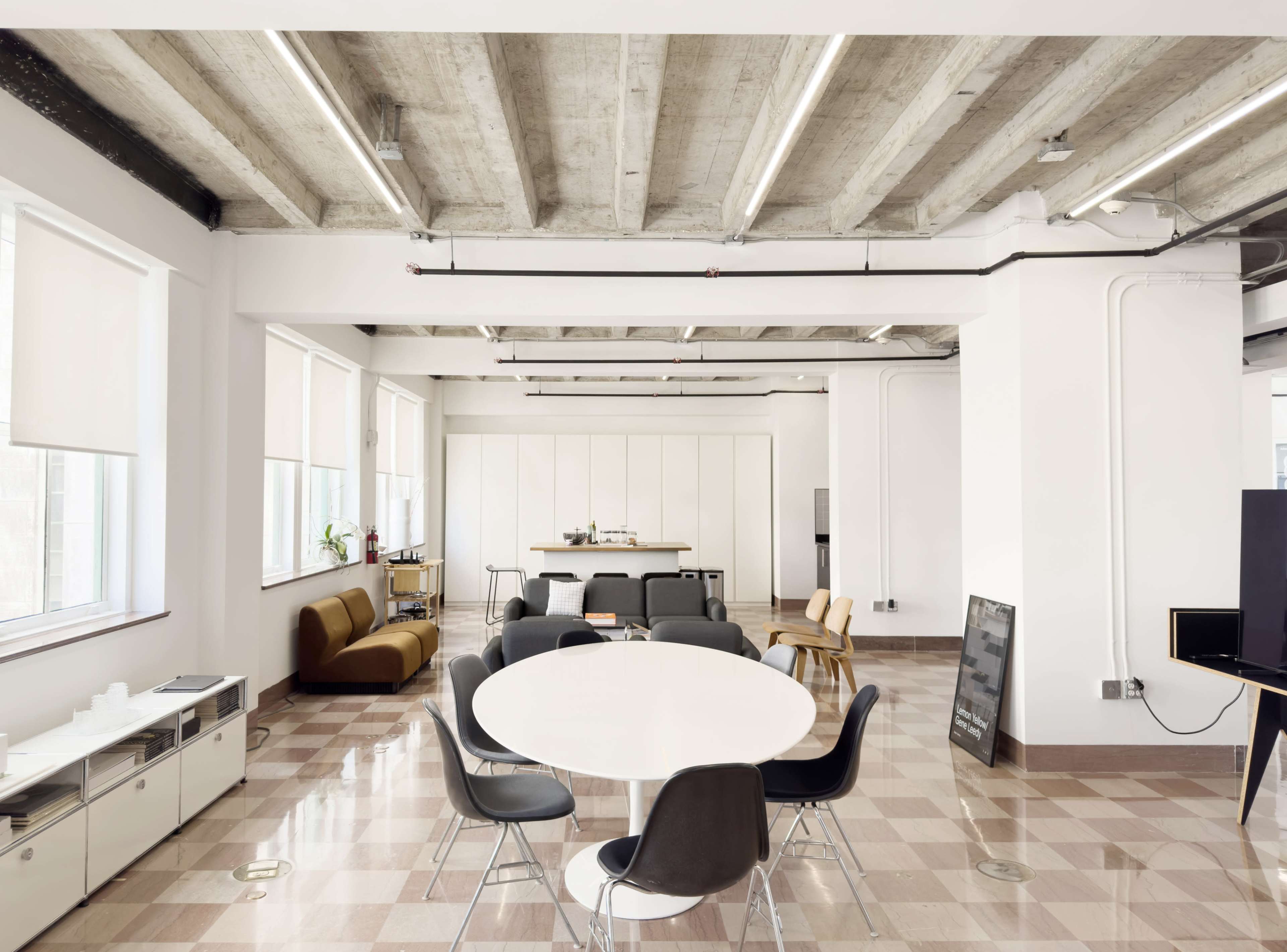 A spacious office area features a round dining table surrounded by black chairs, with a brown couch and modern kitchen cabinetry in the background.