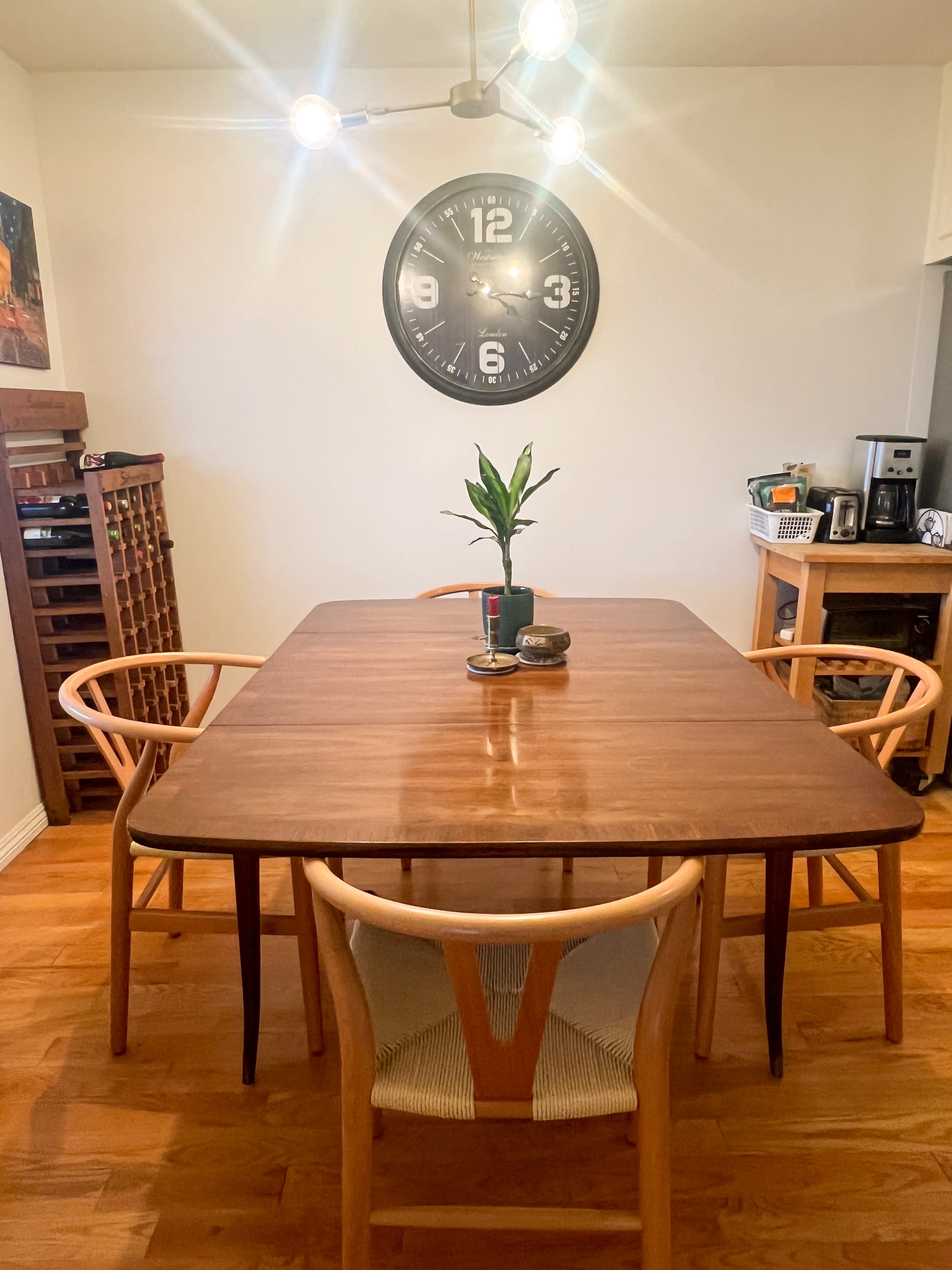 A wooden dining table surrounded by four chairs is set in a well-lit room, with a large clock and a plant on the table.