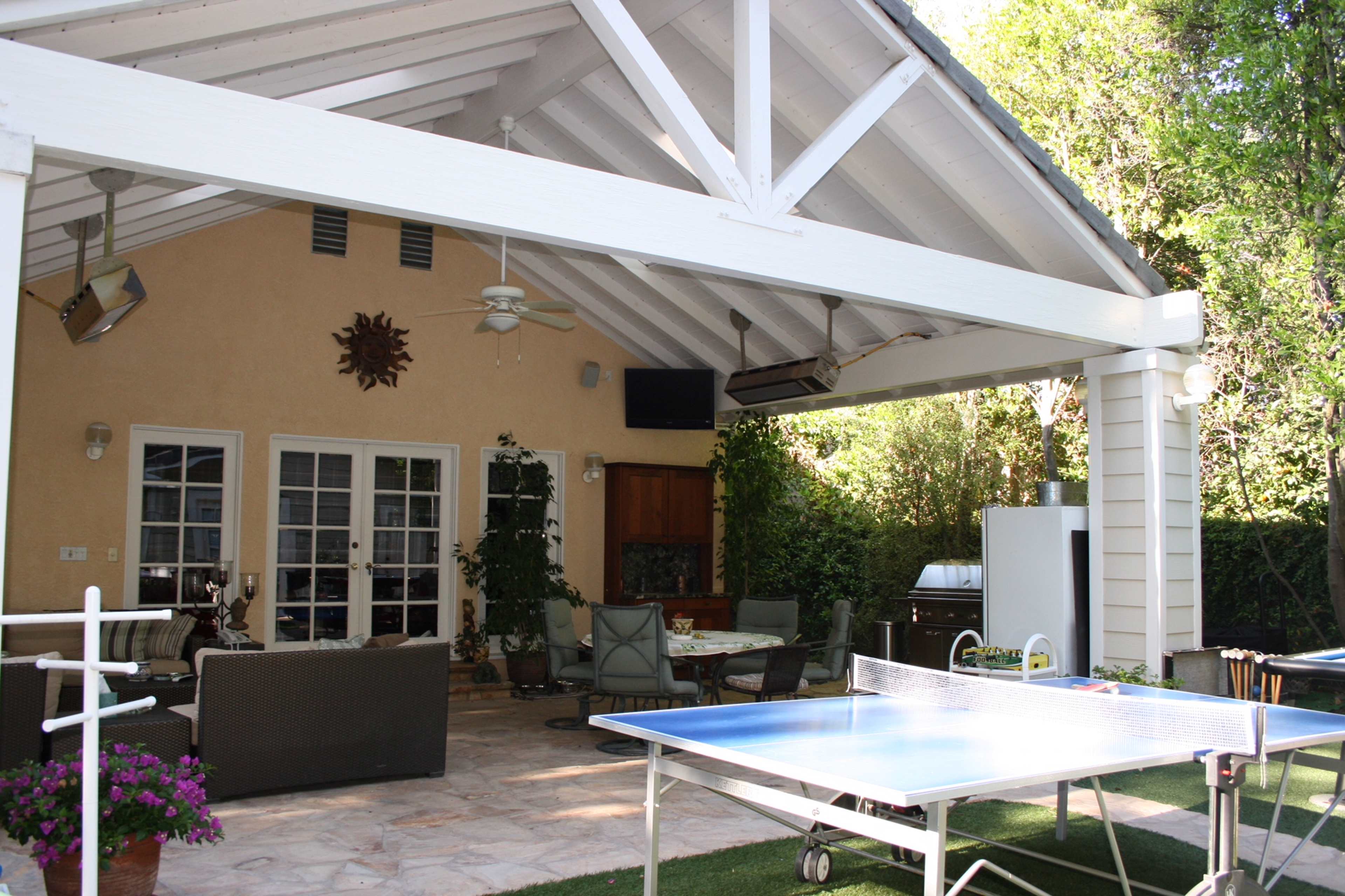 A covered patio features a dining area, a barbecue, and a ping pong table surrounded by greenery.