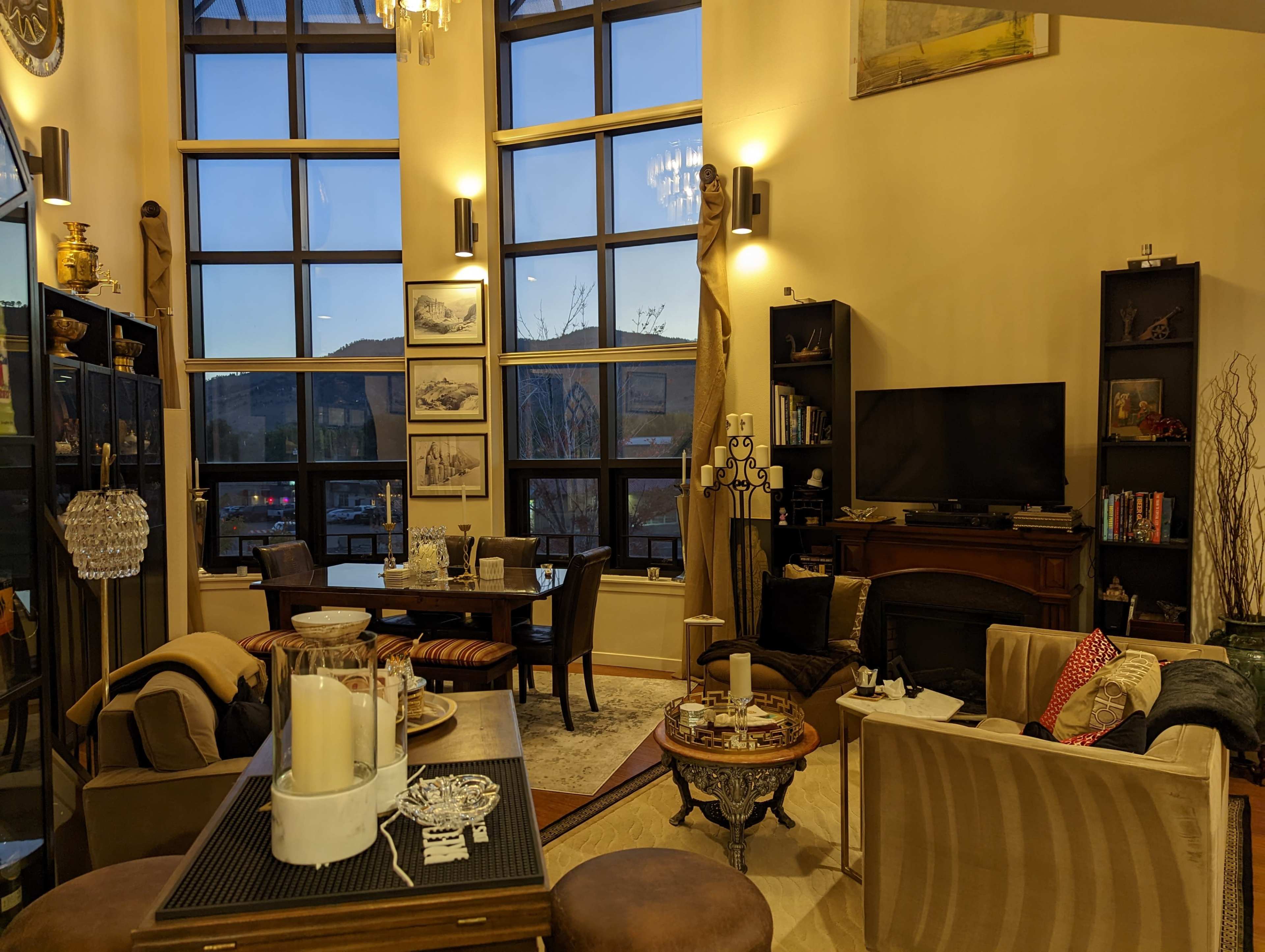 The image shows a modern living room with large windows, a dining area, and various furniture pieces including a couch, chairs, and a television.