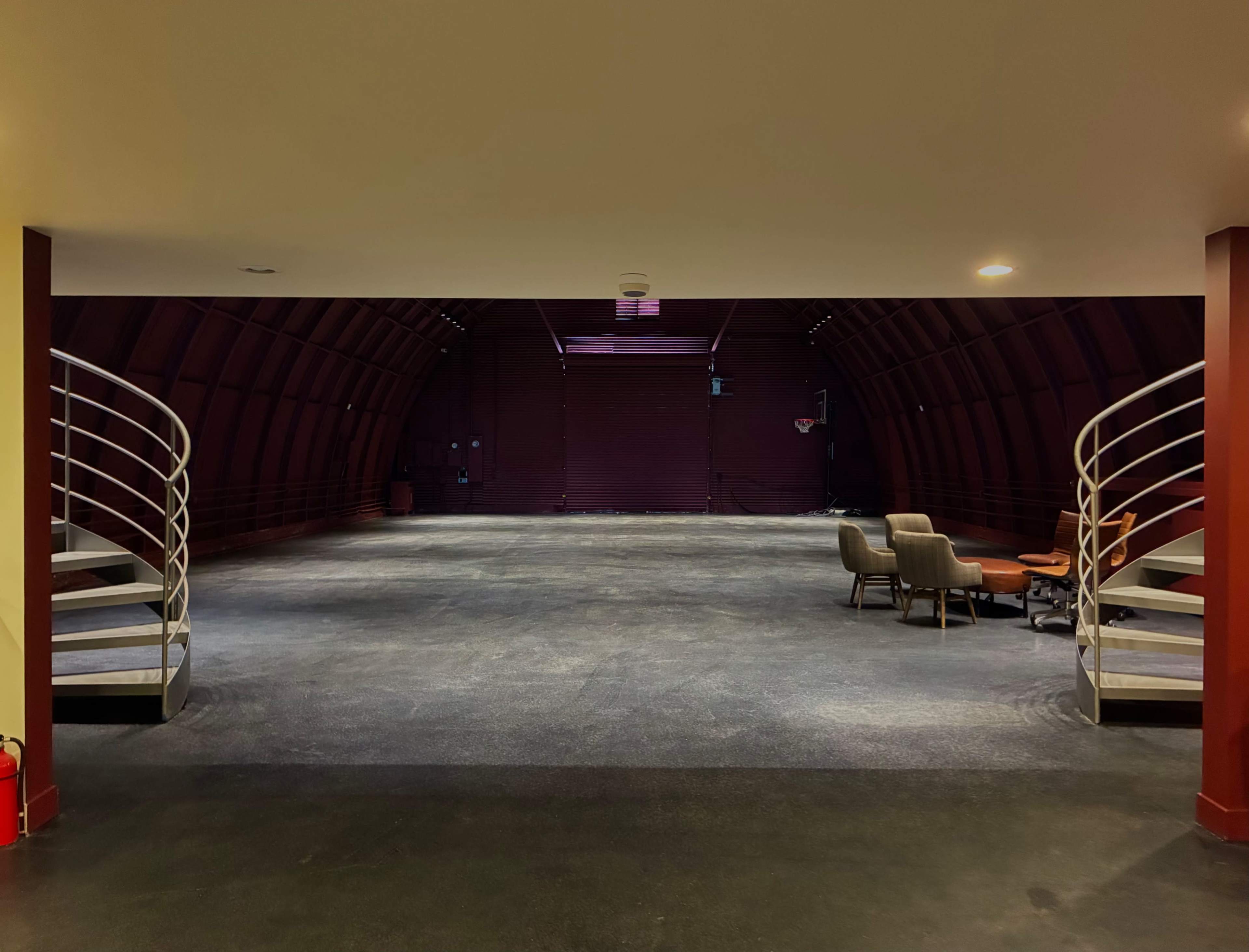The image shows a spacious, empty interior of a curved structure with stairs on either side leading to a lower level, furnished with two chairs and a small table.