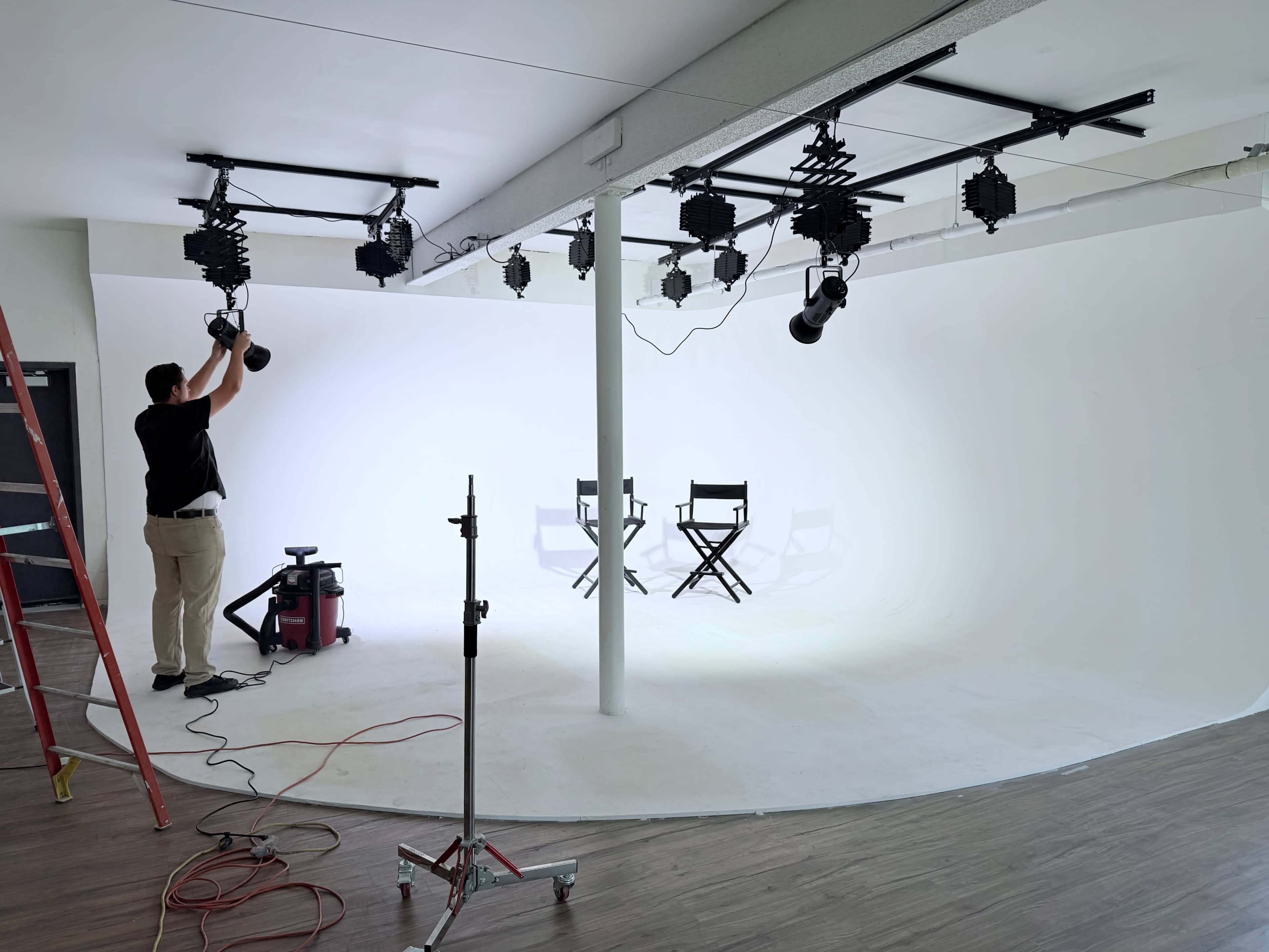 A person adjusts lighting equipment in a photography studio featuring a seamless white backdrop and two empty director's chairs.