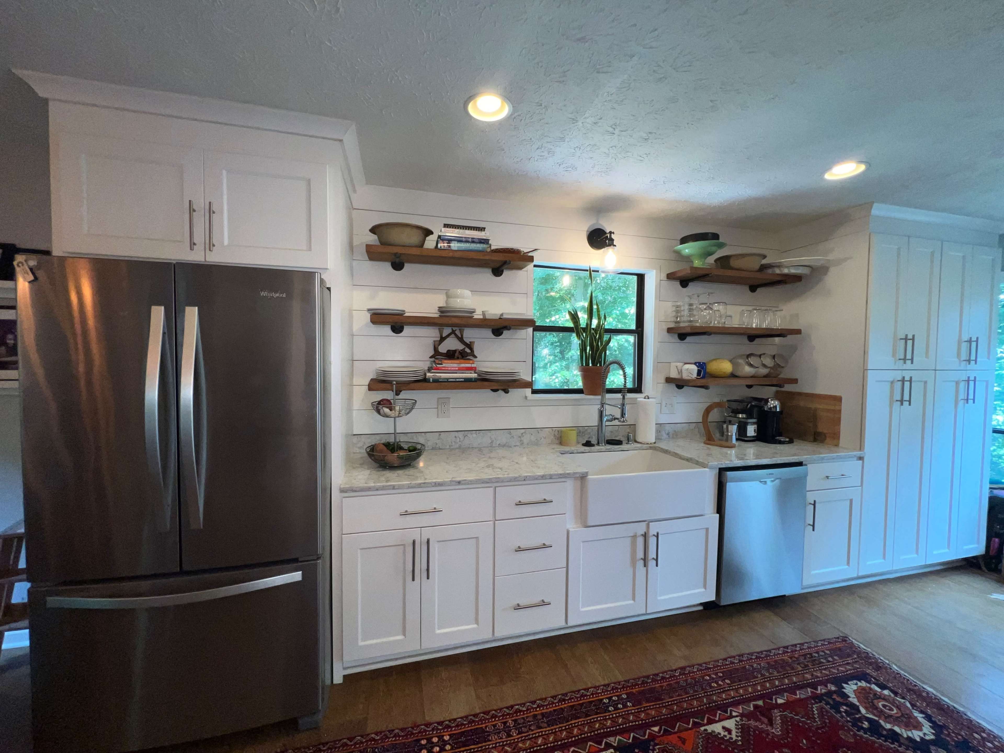 A modern kitchen with white cabinetry, a farmhouse sink, stainless steel appliances, and open shelves displaying dishware and decorative items.