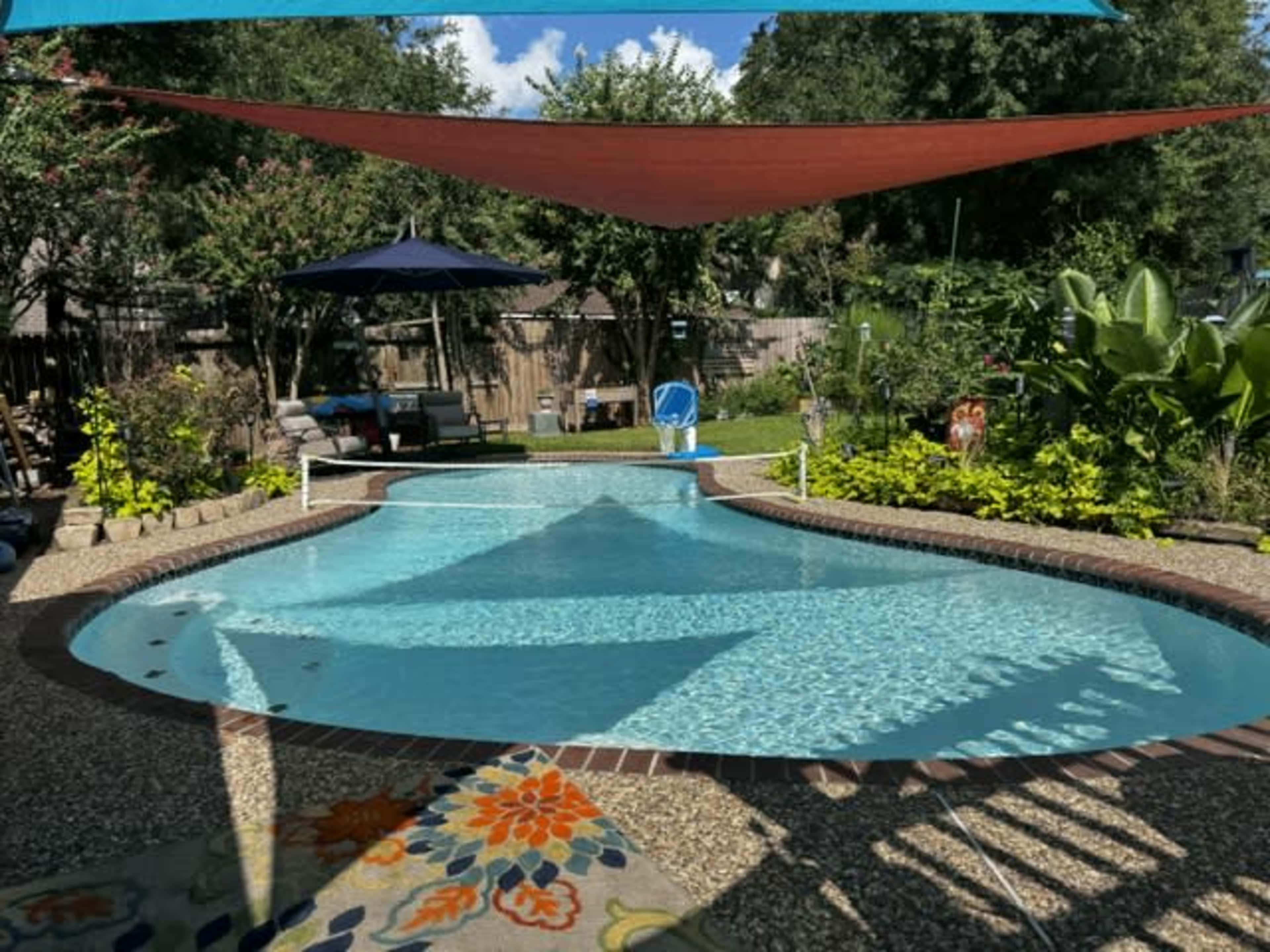 The image shows a swimming pool surrounded by lush greenery, lounge chairs, and shade sails in a backyard setting.