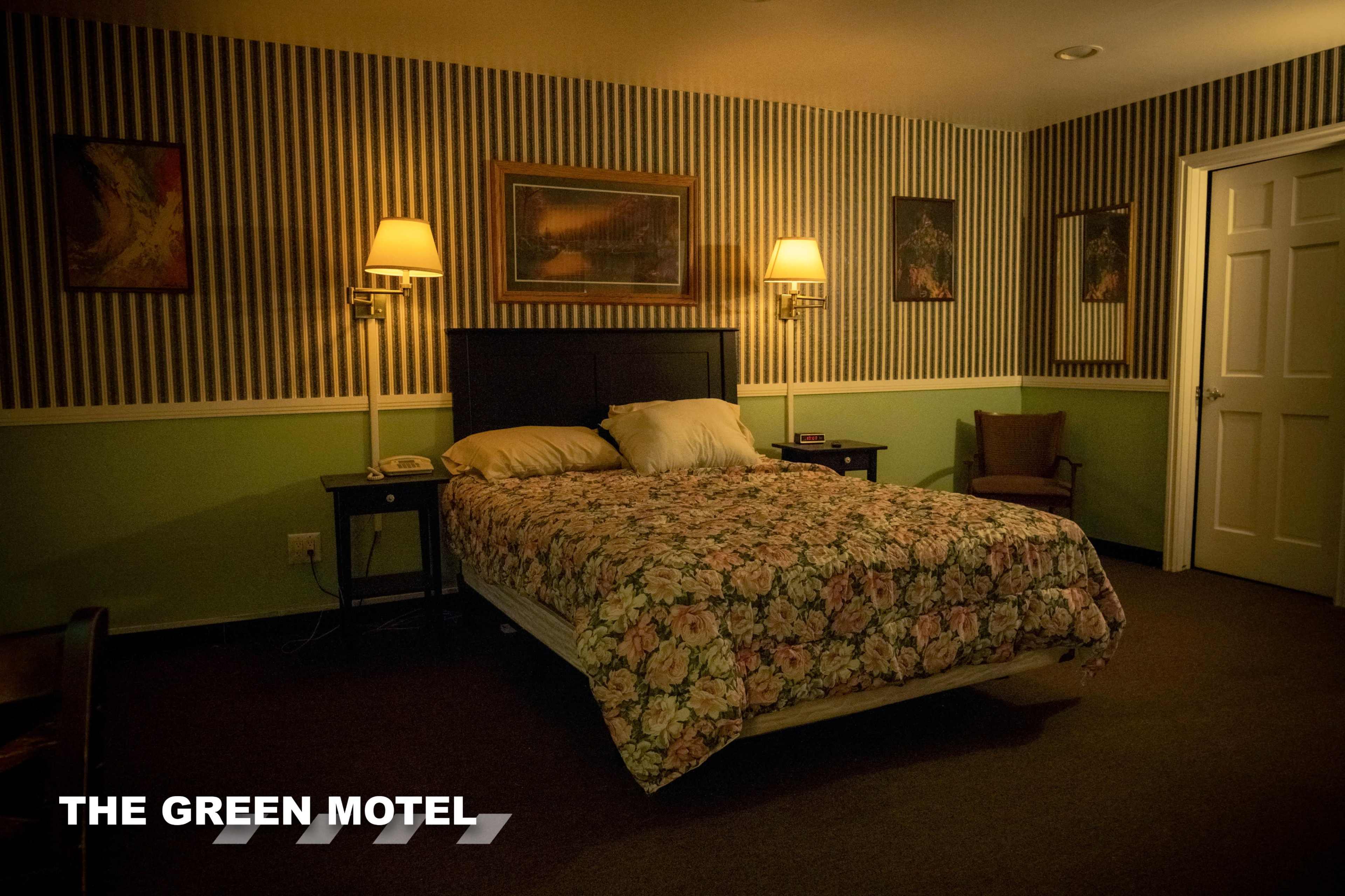 The image shows a motel room with a double bed covered in floral bedding, two wall lamps, and a chair beside a door.