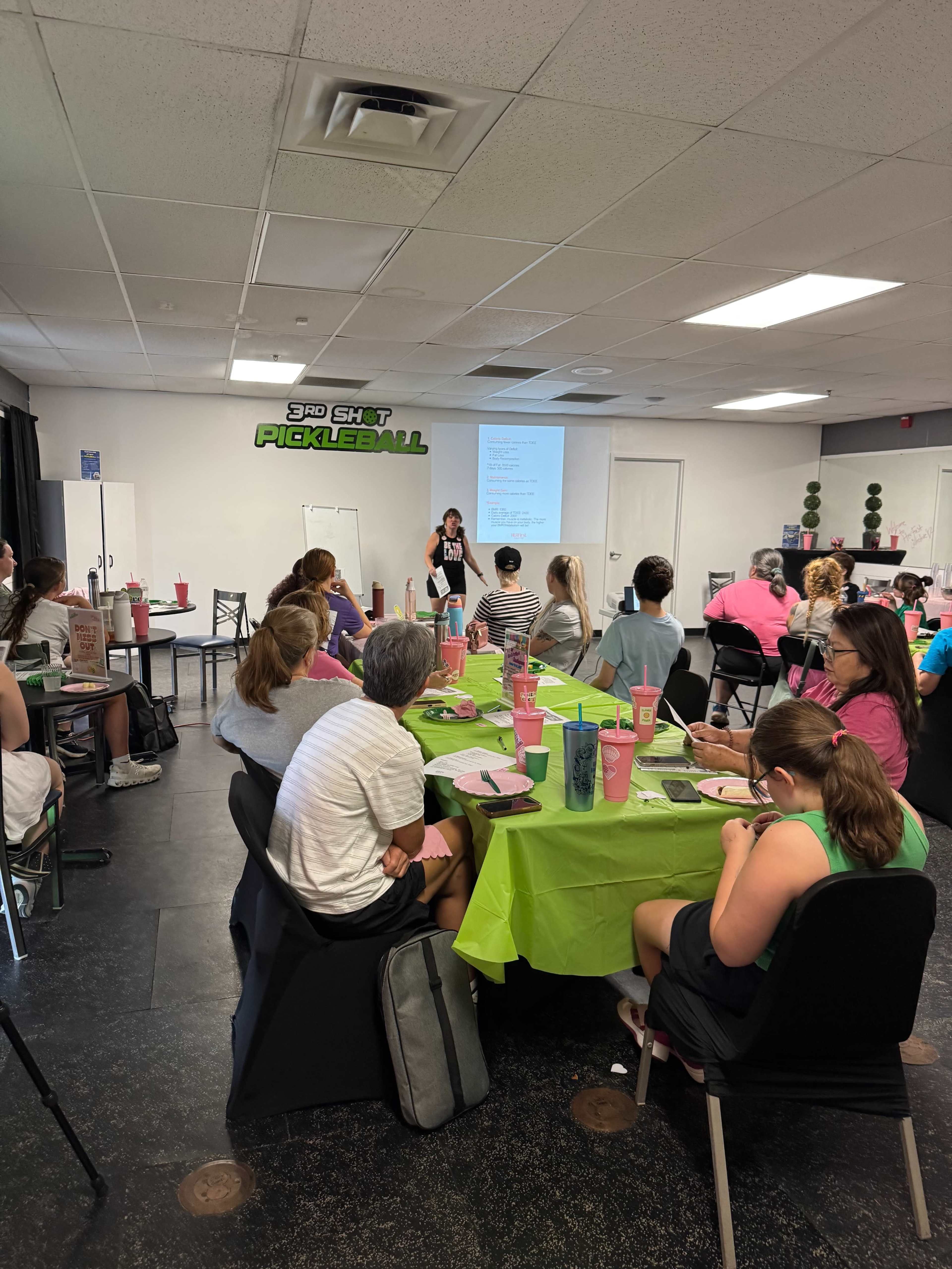A group of people sits at tables covered in green tablecloths, attentively listening to a speaker in a room decorated for a presentation about pickleball.