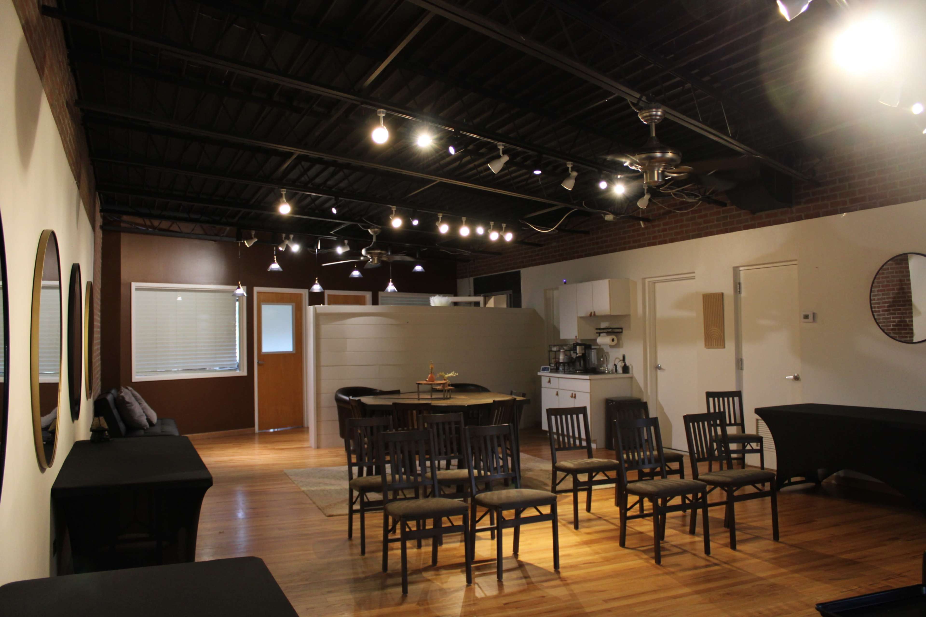 An indoor space with wooden floors, several chairs arranged in rows, a kitchenette area, and soft lighting from ceiling fixtures.