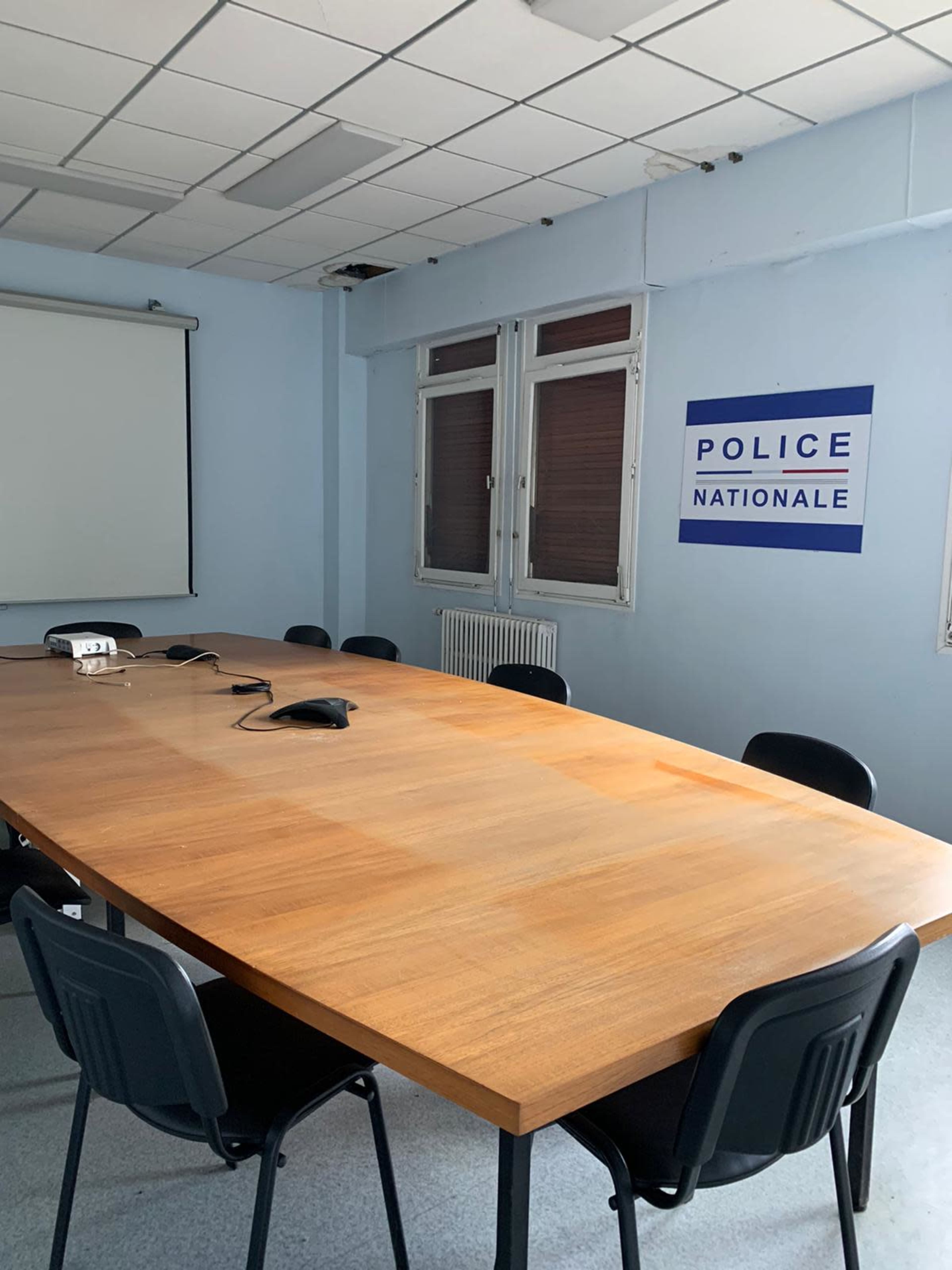 A conference room with a large wooden table surrounded by black chairs and a wall displaying a "POLICE NATIONALE" sign.