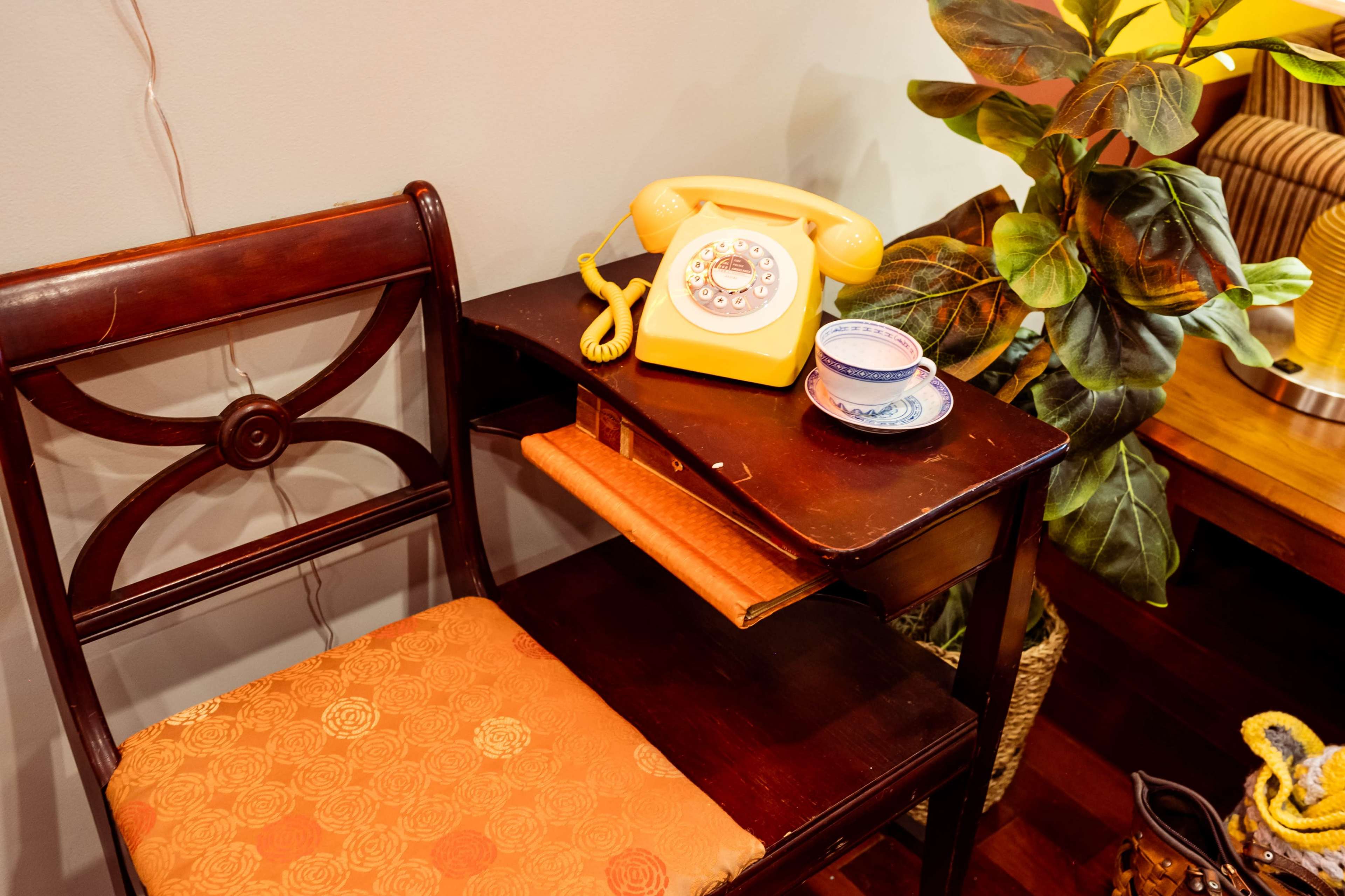 A wooden chair is placed next to a small table featuring a yellow rotary telephone, a teacup, and a potted plant.