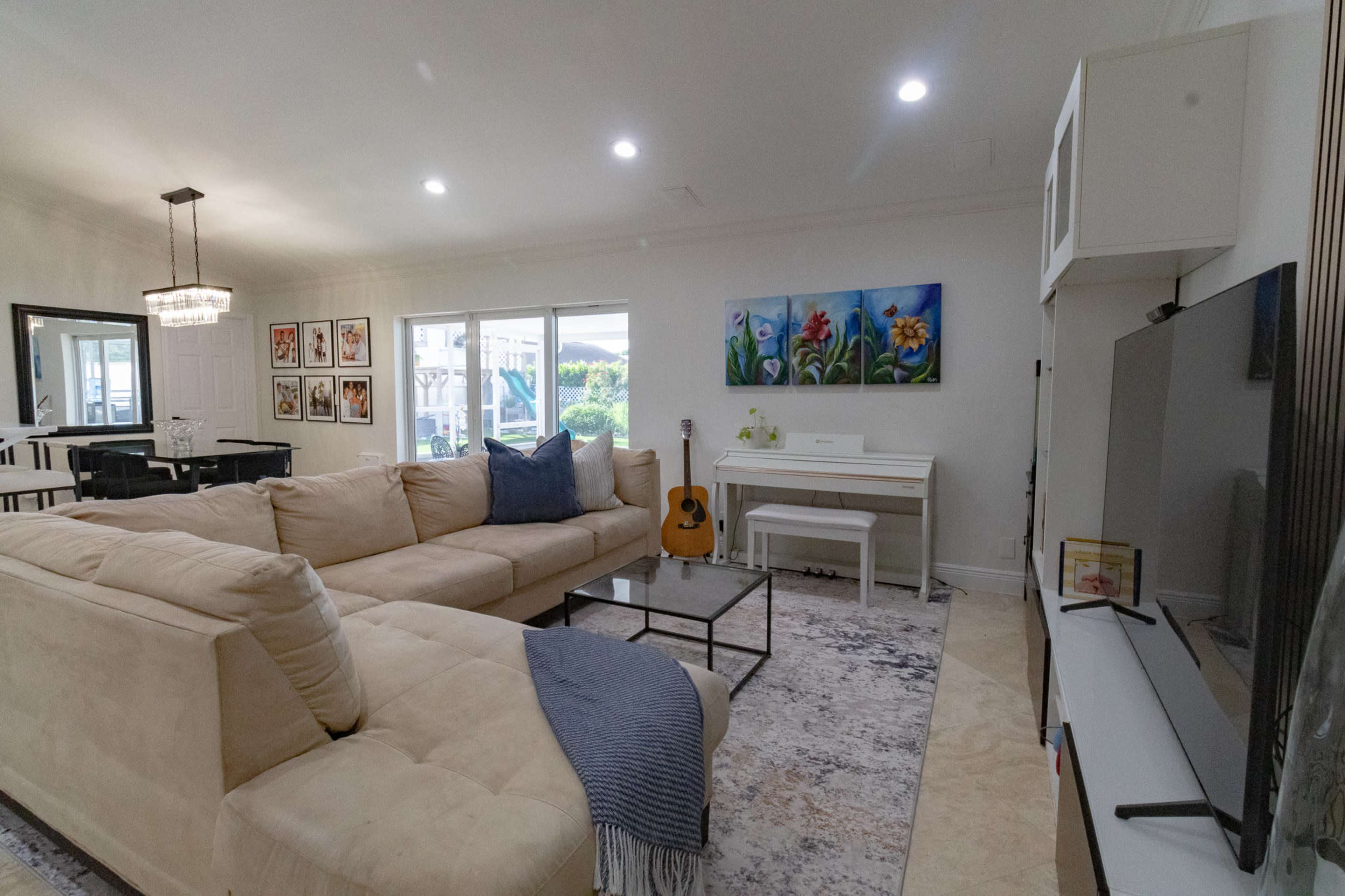 A spacious living room features a large beige sectional sofa, a glass coffee table, a piano, and artwork on the walls, with natural light streaming through the windows.