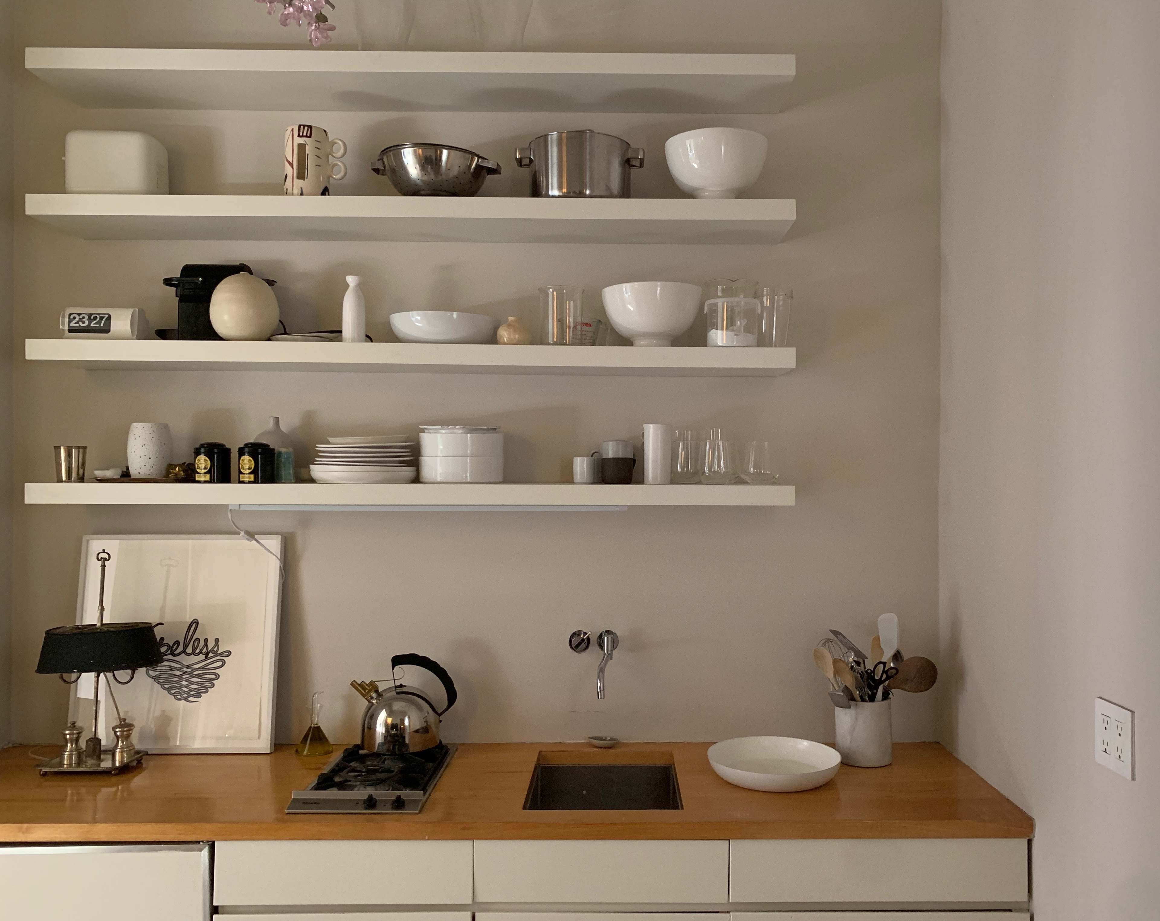 A minimalist kitchen with wooden countertops, white shelves displaying various kitchenware, and a stove with a kettle.