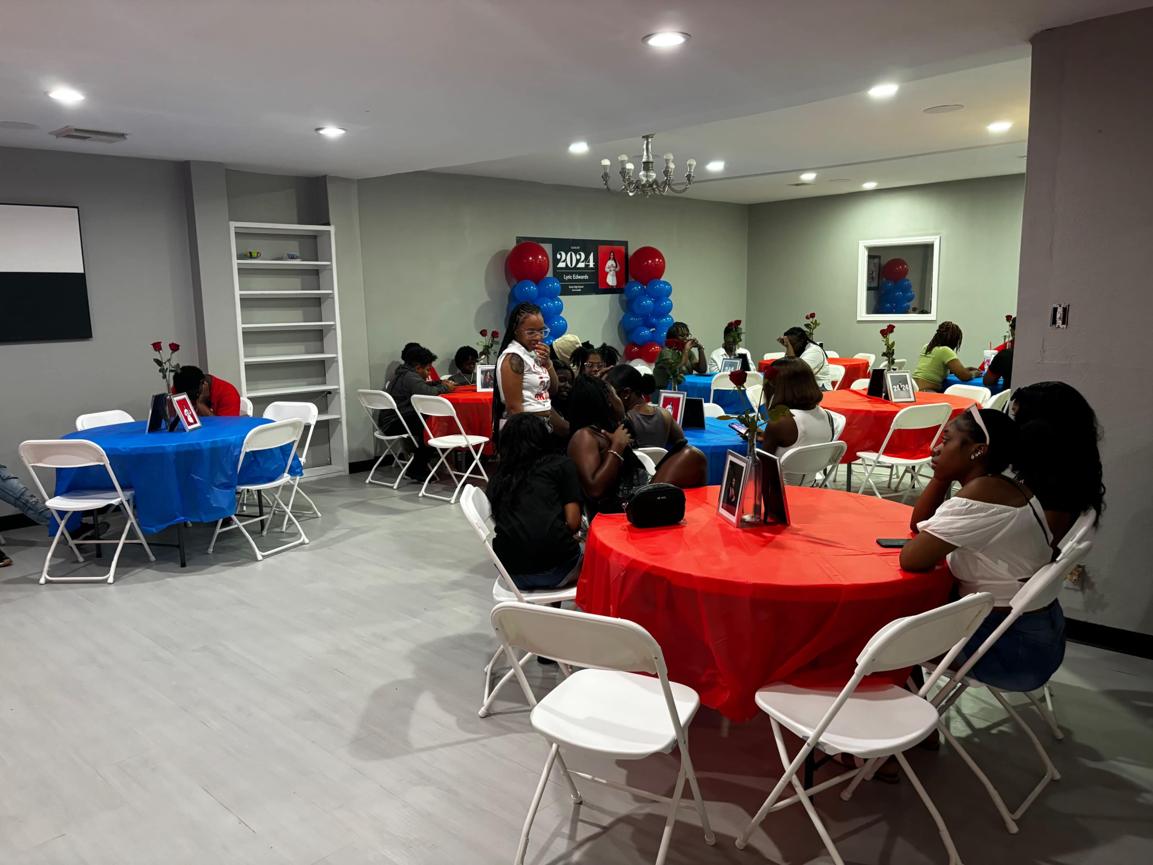 A group of people sits at red and blue table settings in a decorated room, with a banner in the background indicating the year 2024.