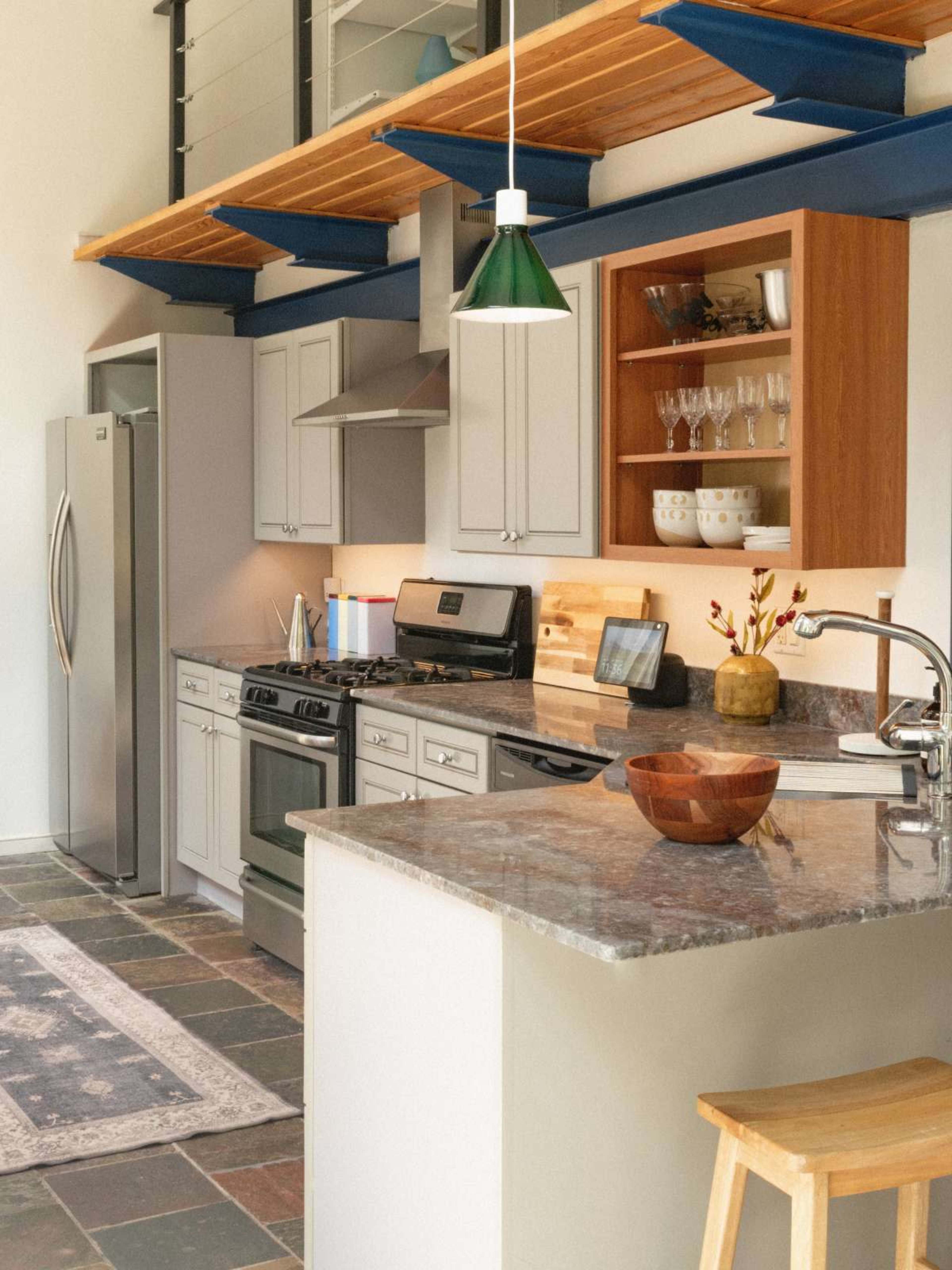 The image shows a modern kitchen with grey cabinets, stainless steel appliances, a granite countertop, and wooden shelving above.