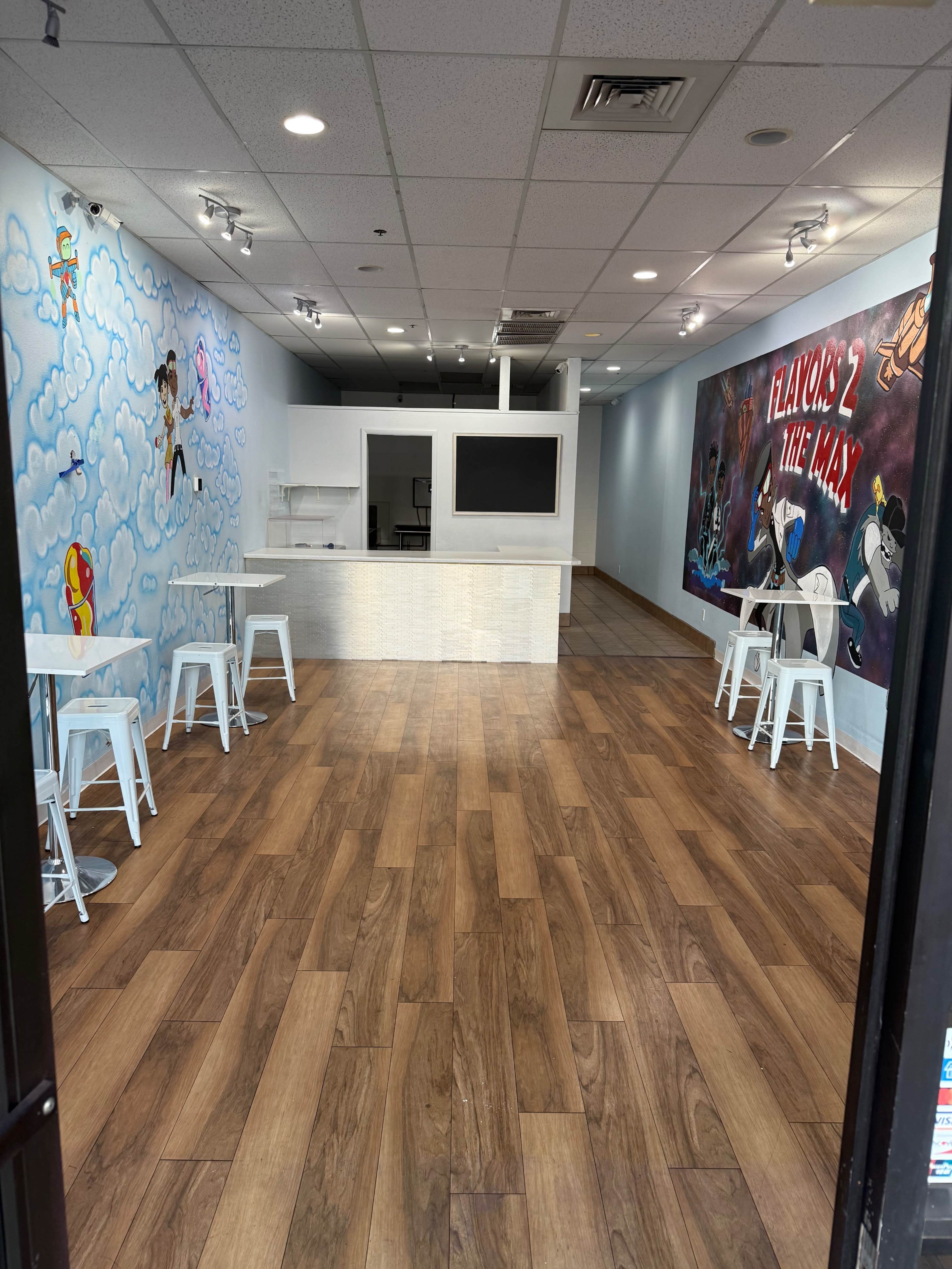 The image shows a modern, empty café with wooden flooring, white tables, and colorful wall murals depicting animated characters and text.