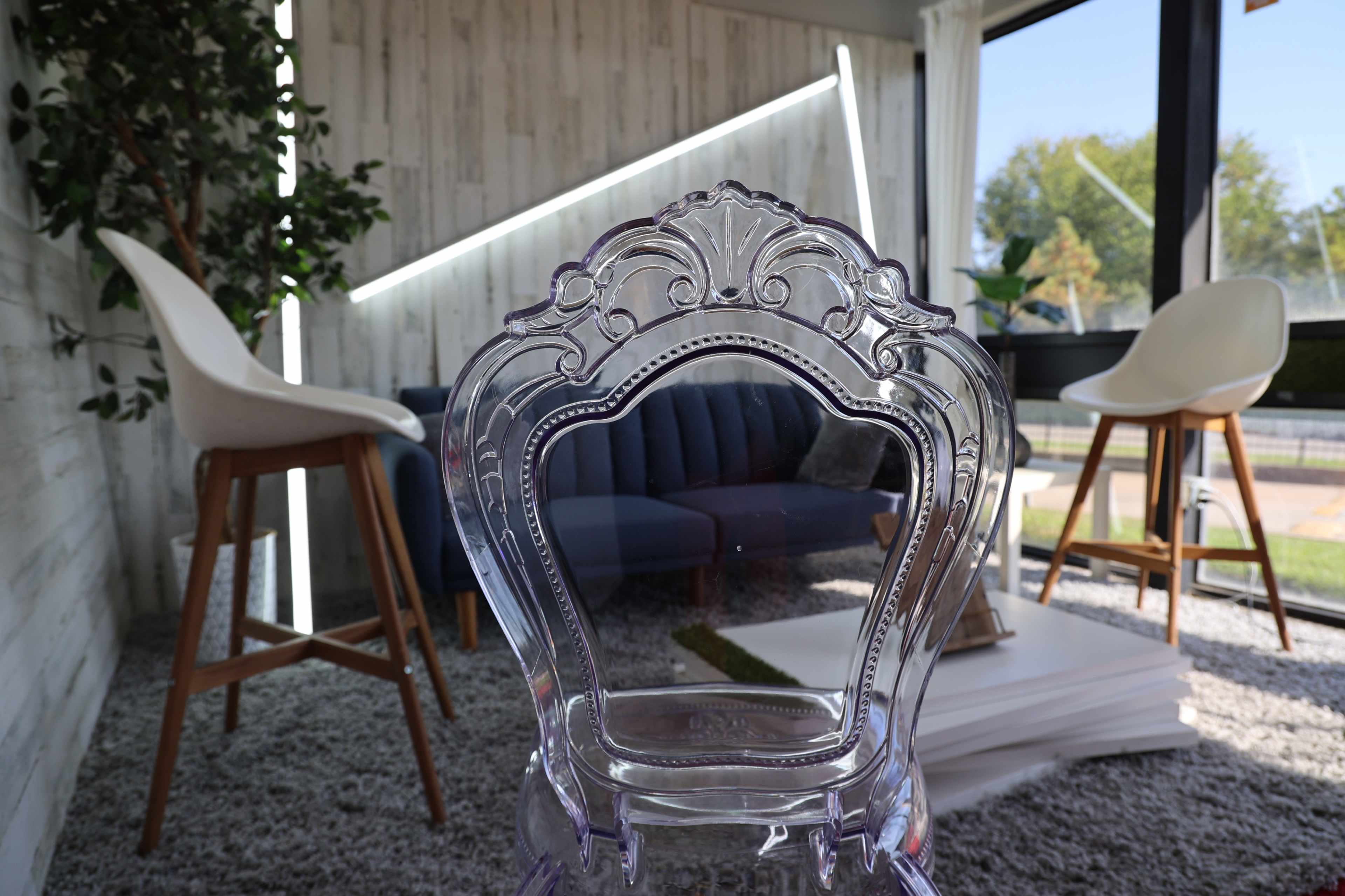 A transparent chair is positioned in the foreground, facing a cozy seating area with a blue couch and two tall stools in a well-lit room.