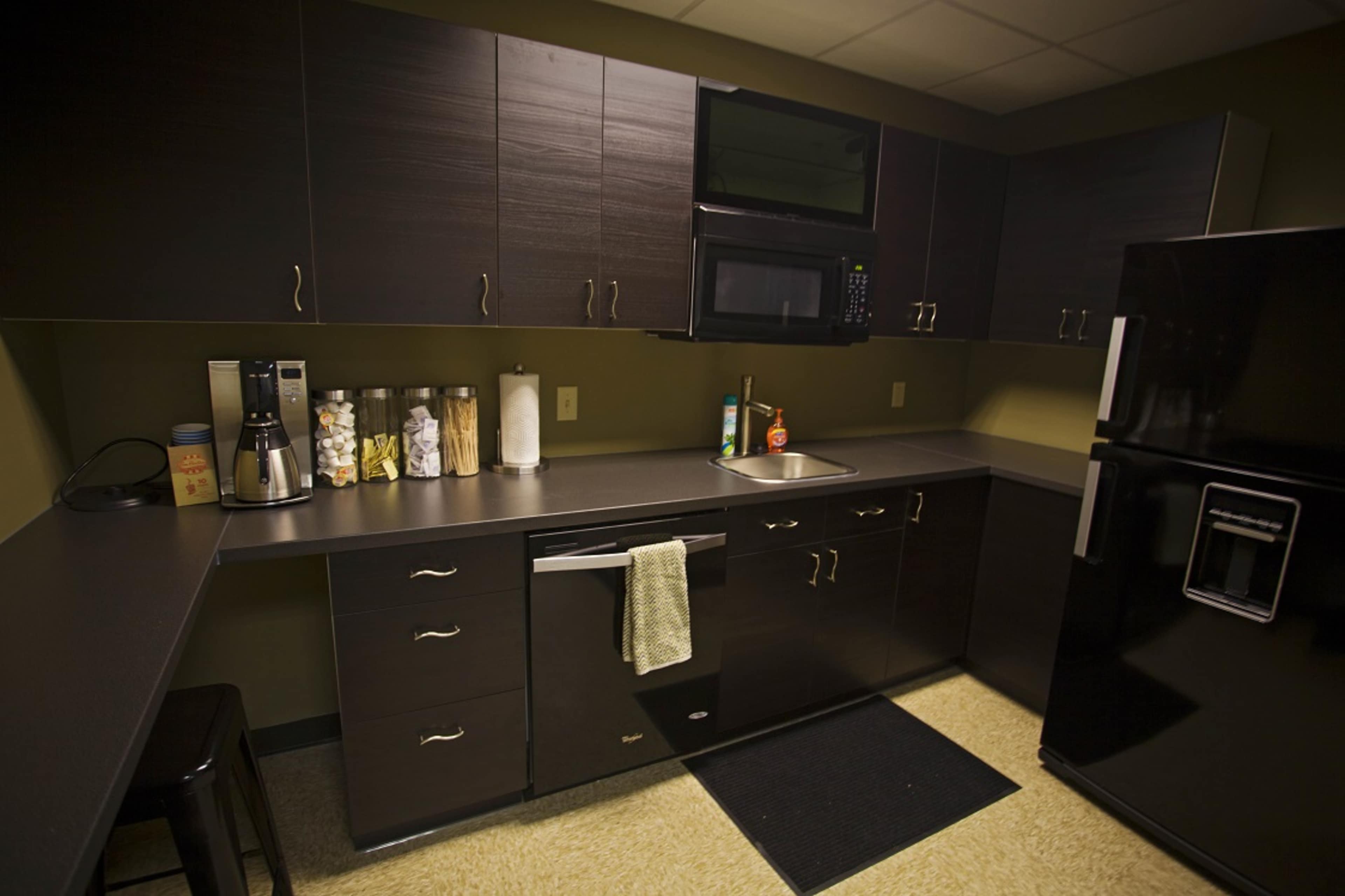 The image shows a modern kitchen with dark cabinetry, a countertop, a microwave, a coffee maker, and a refrigerator.