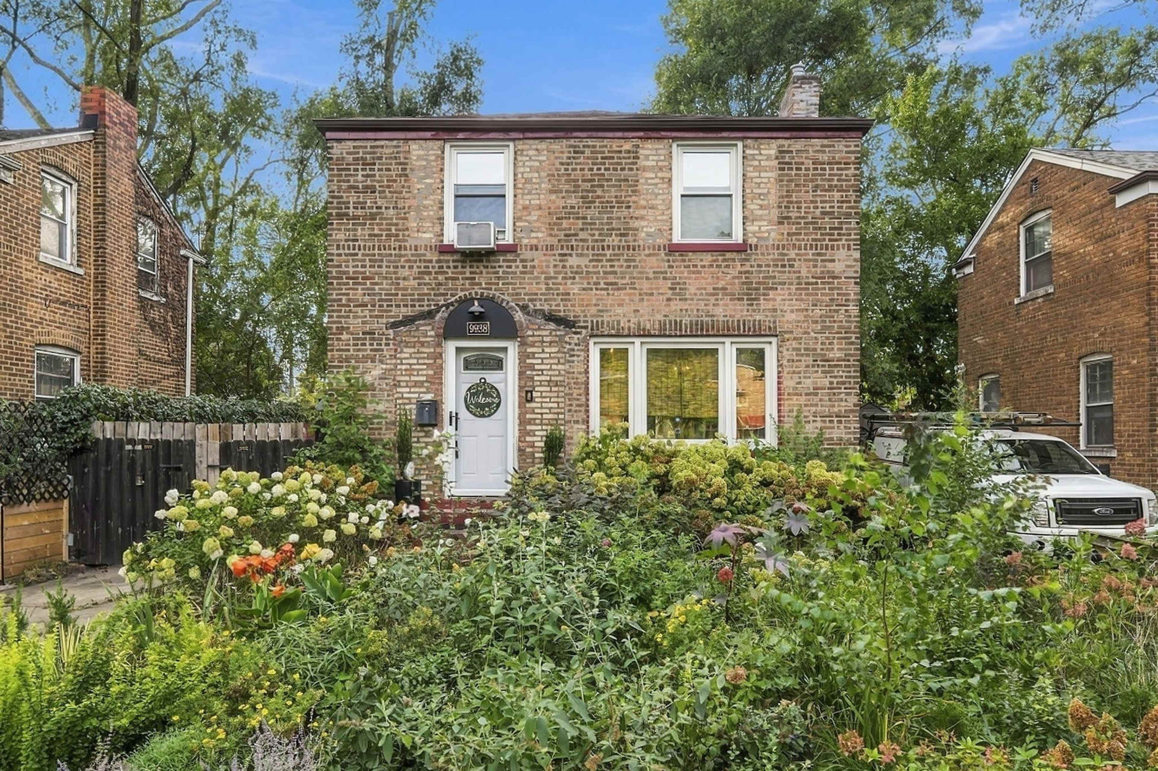 A two-story brick house surrounded by a vibrant garden filled with various plants and flowers.