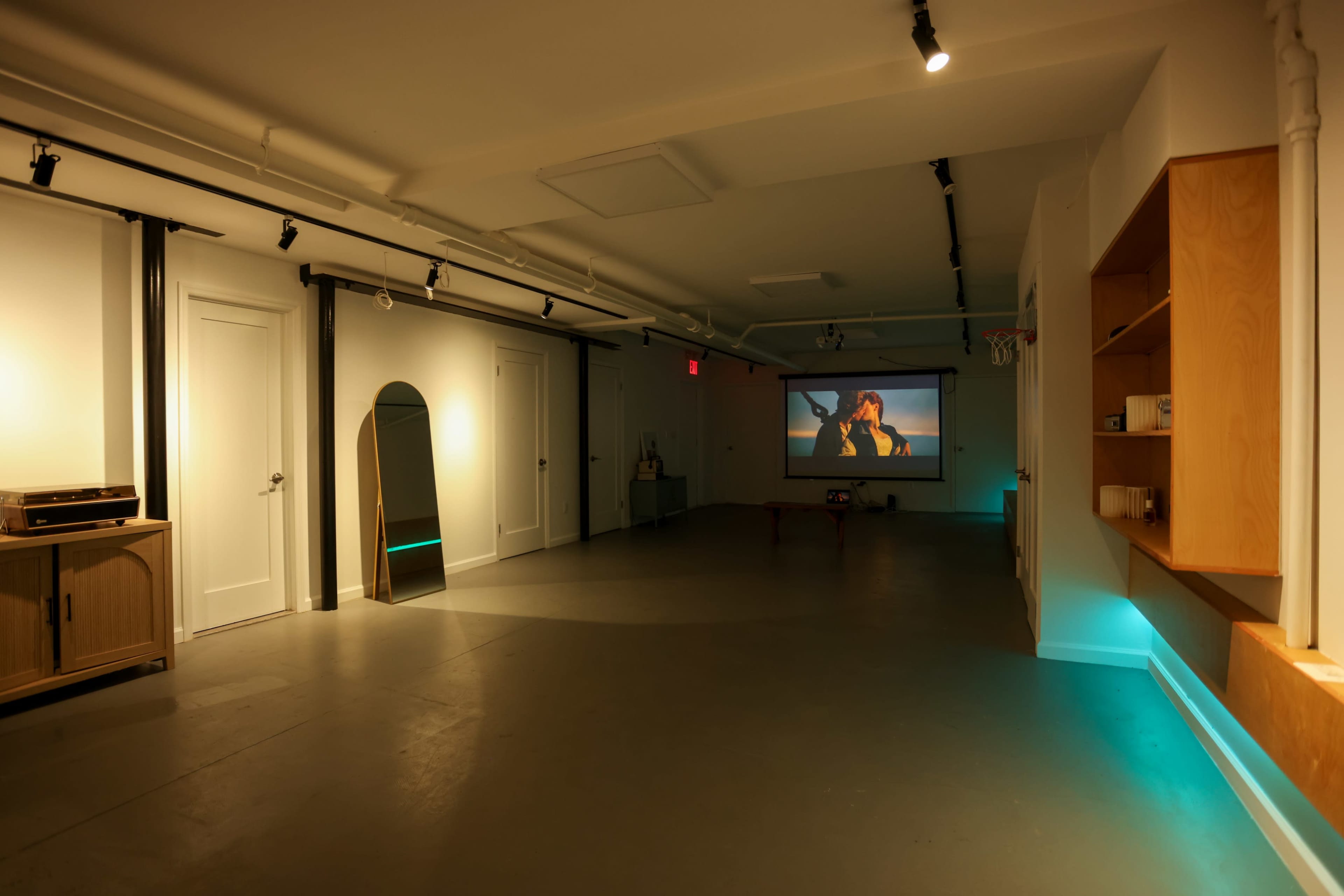 A spacious, well-lit room features a large projection screen displaying a film, with minimalist decor including a mirror and shelving along the walls.