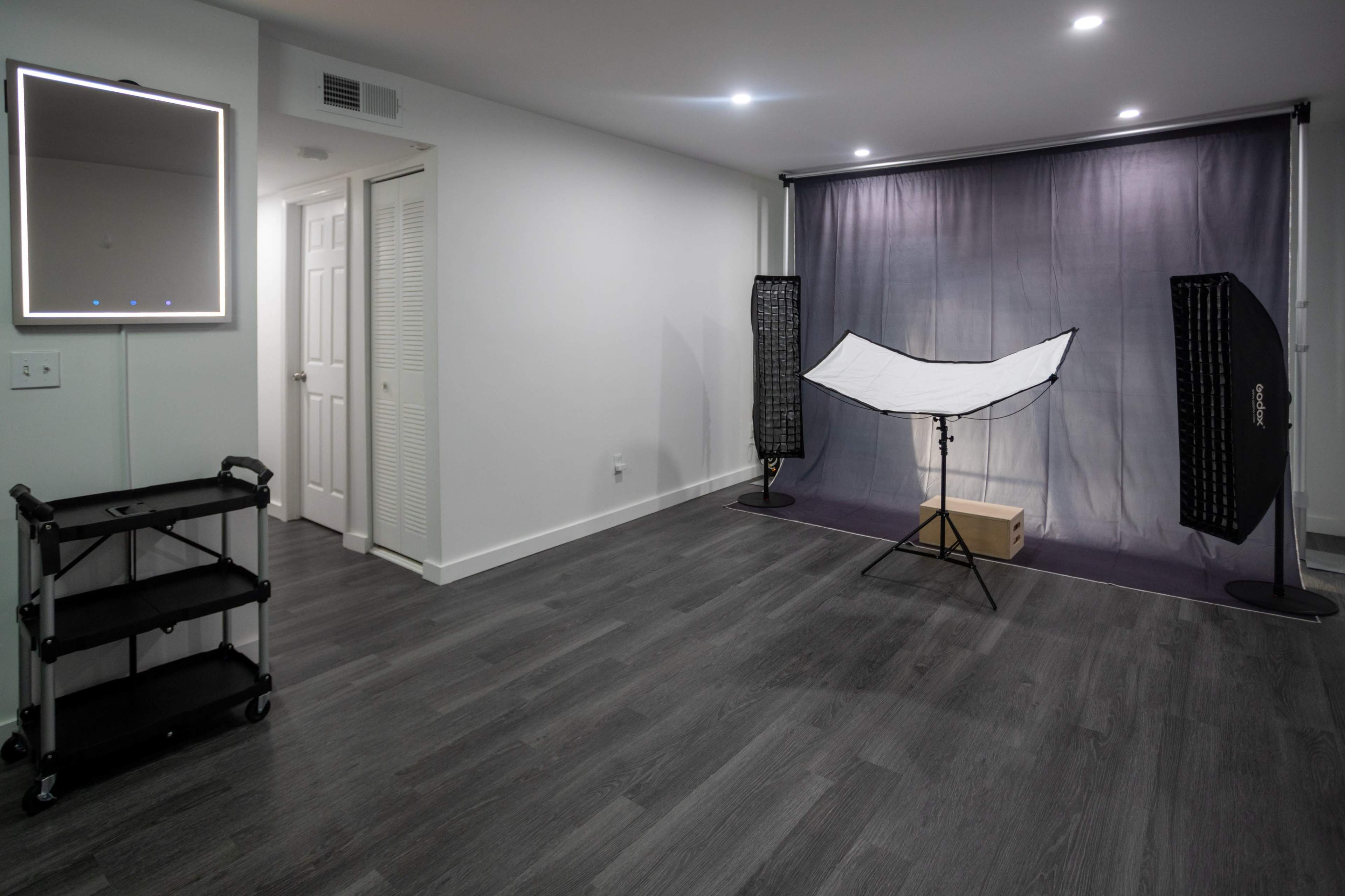 The image shows a well-lit room featuring a backdrop setup with softboxes, a mirror on the wall, and a black cart in the corner.