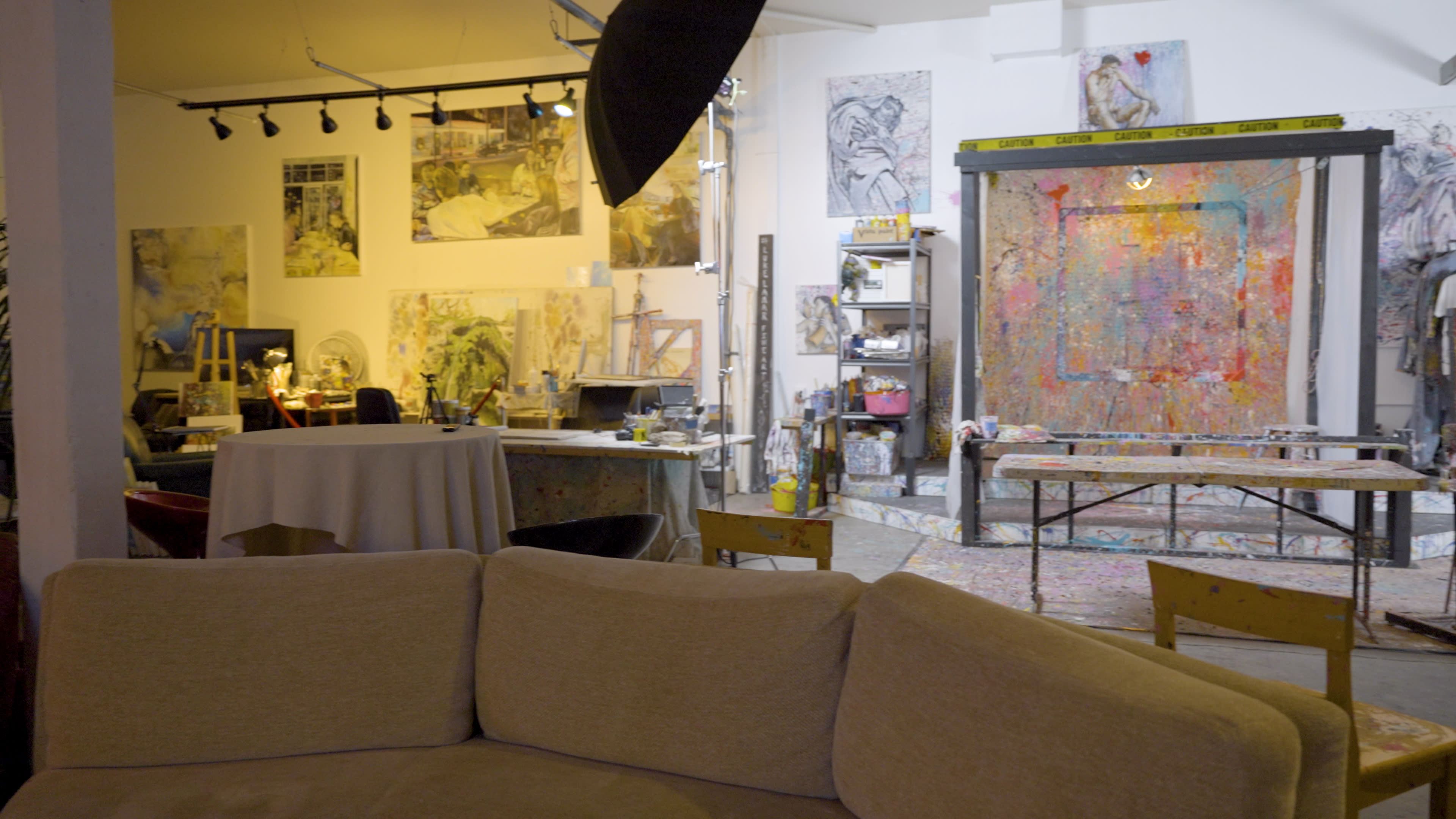 An art studio with various paintings on the walls, a large colorful canvas at the back, and furniture including a couch and tables scattered throughout the space.