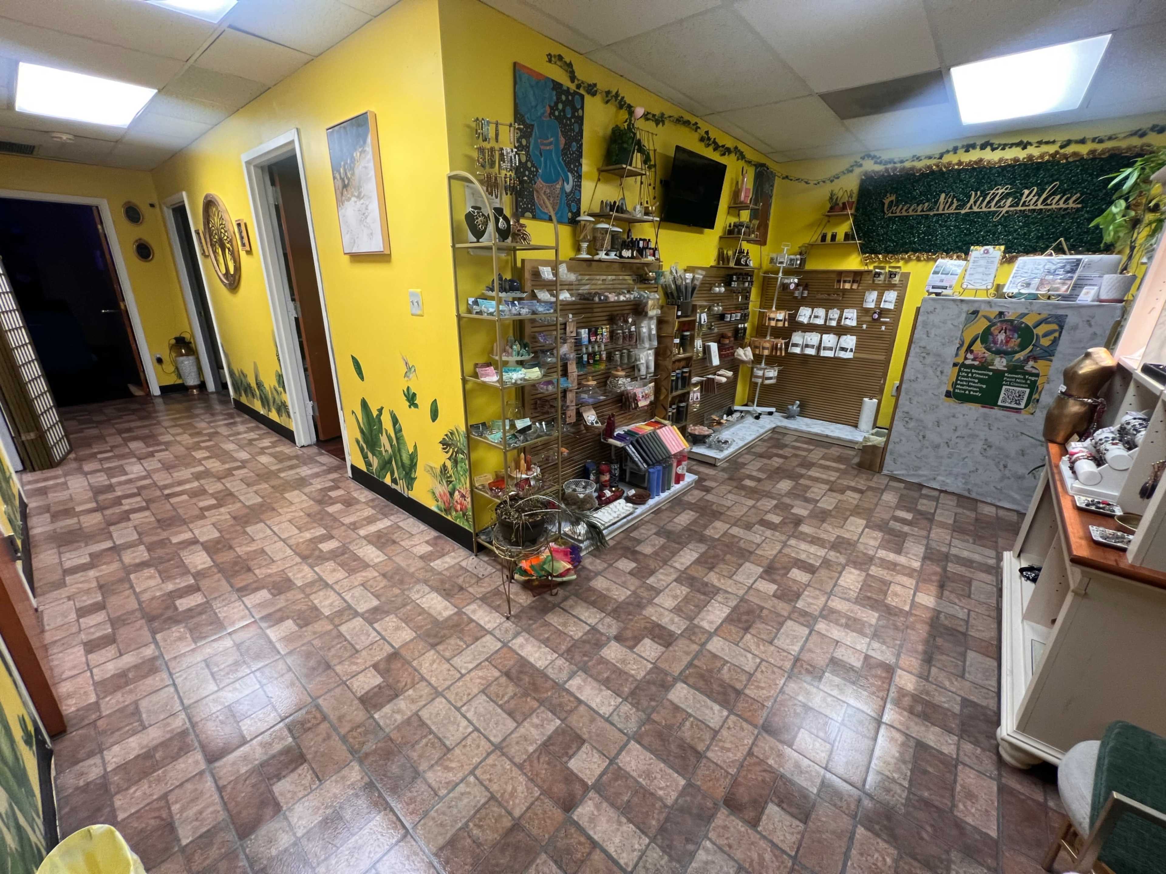 The image shows a vibrant yellow room with tiled flooring, featuring shelves filled with various products and a wall decorated with green foliage artwork.