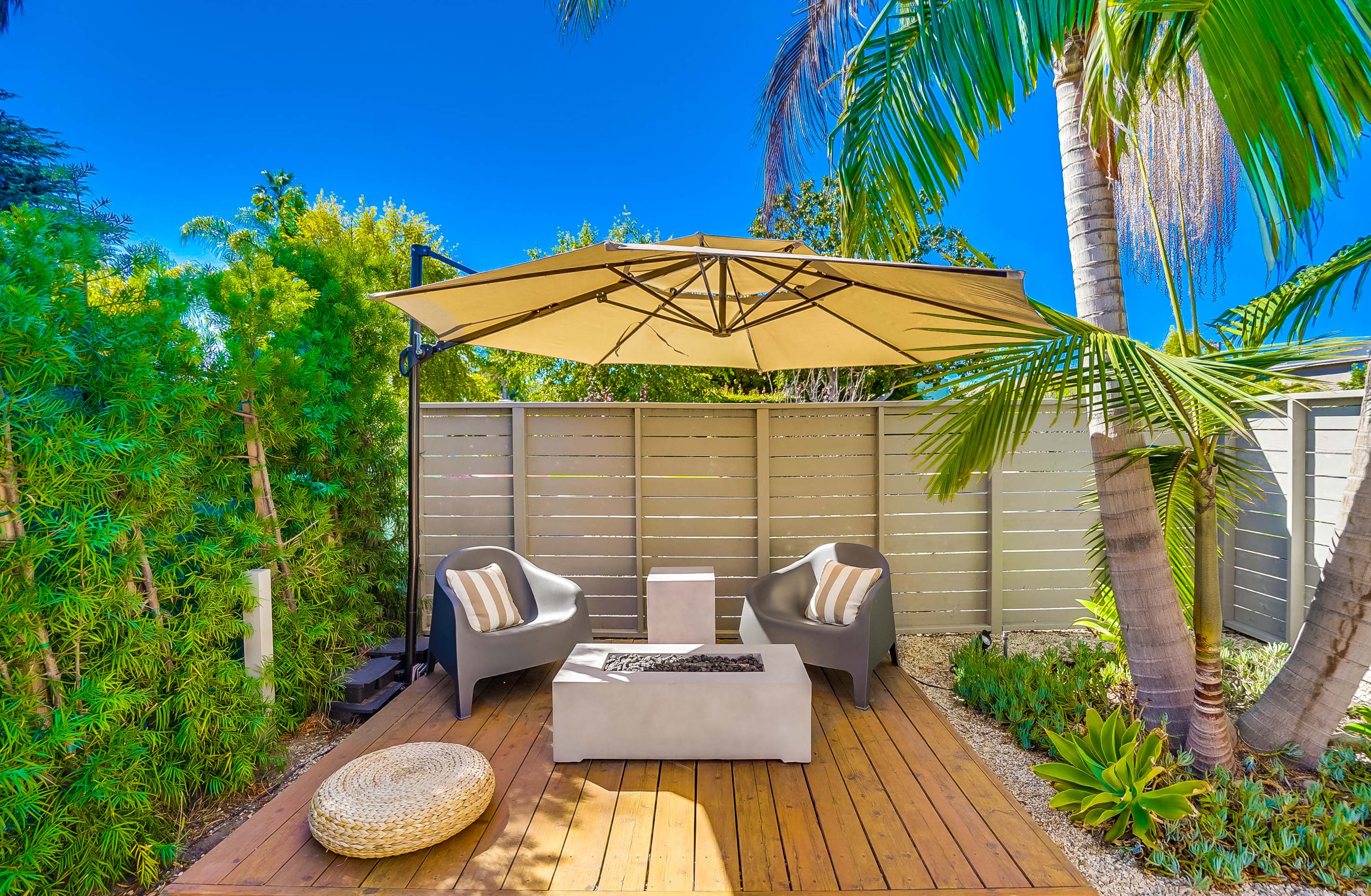 The image shows a patio area featuring two gray chairs, a central table with a fire pit, and a large umbrella, surrounded by greenery and a wooden deck.