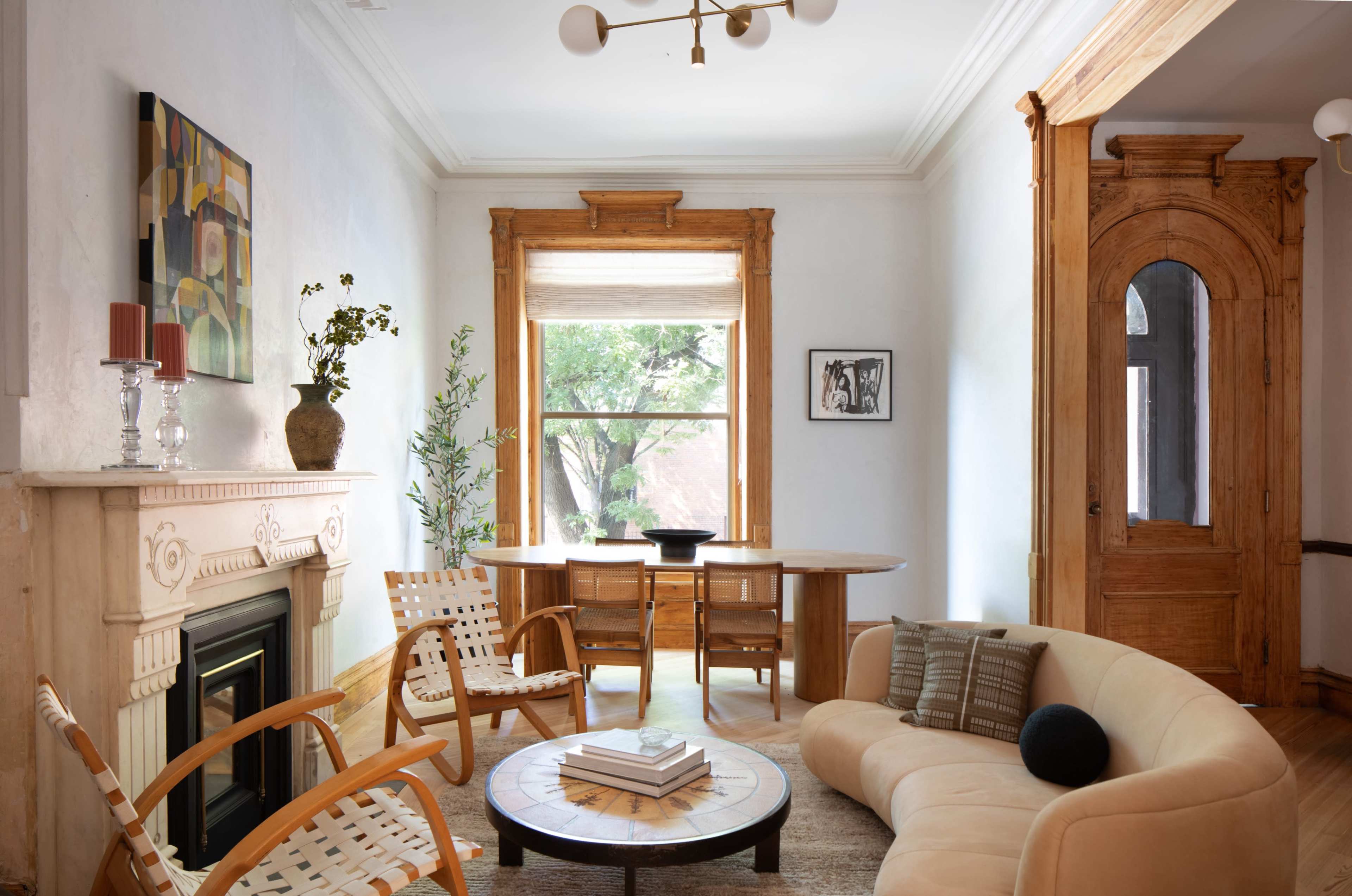 The image shows a cozy living room featuring a modern sofa, wooden armchairs, a round dining table, and large windows framed with classic wooden trim.