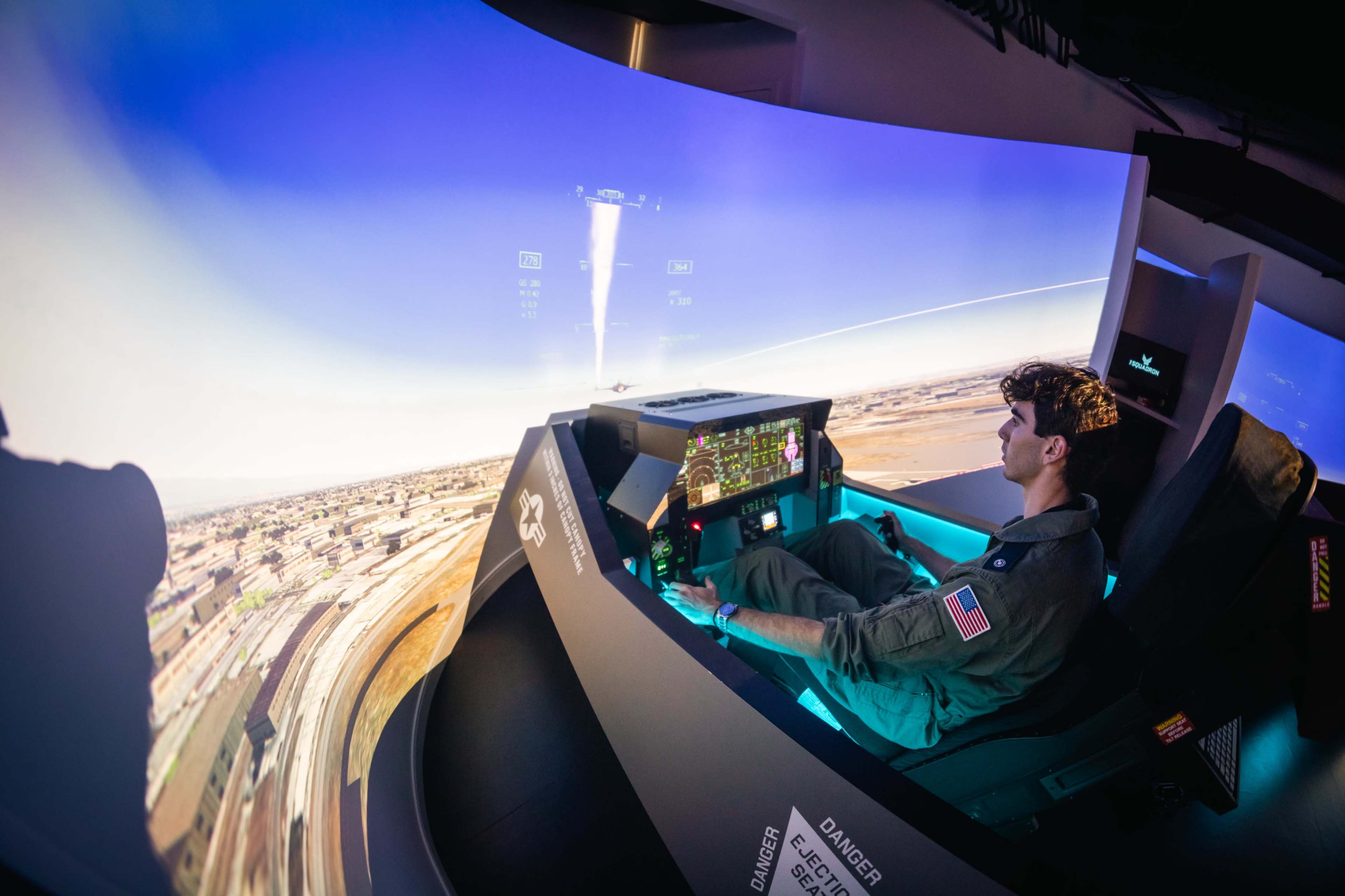 A pilot operates a flight simulator with a panoramic display of a landscape in front of him.
