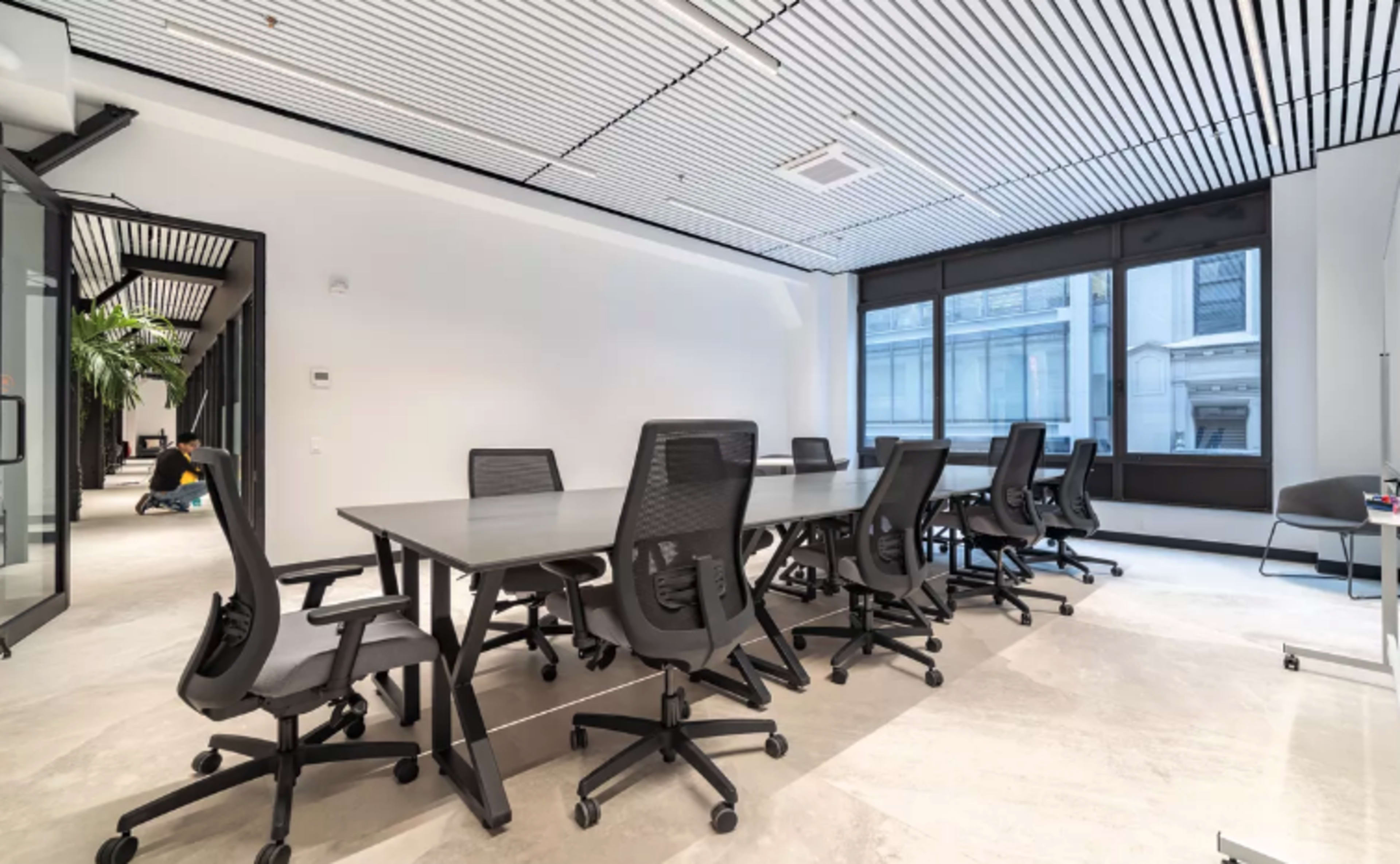 A modern conference room features a large table surrounded by several office chairs and large windows providing natural light.