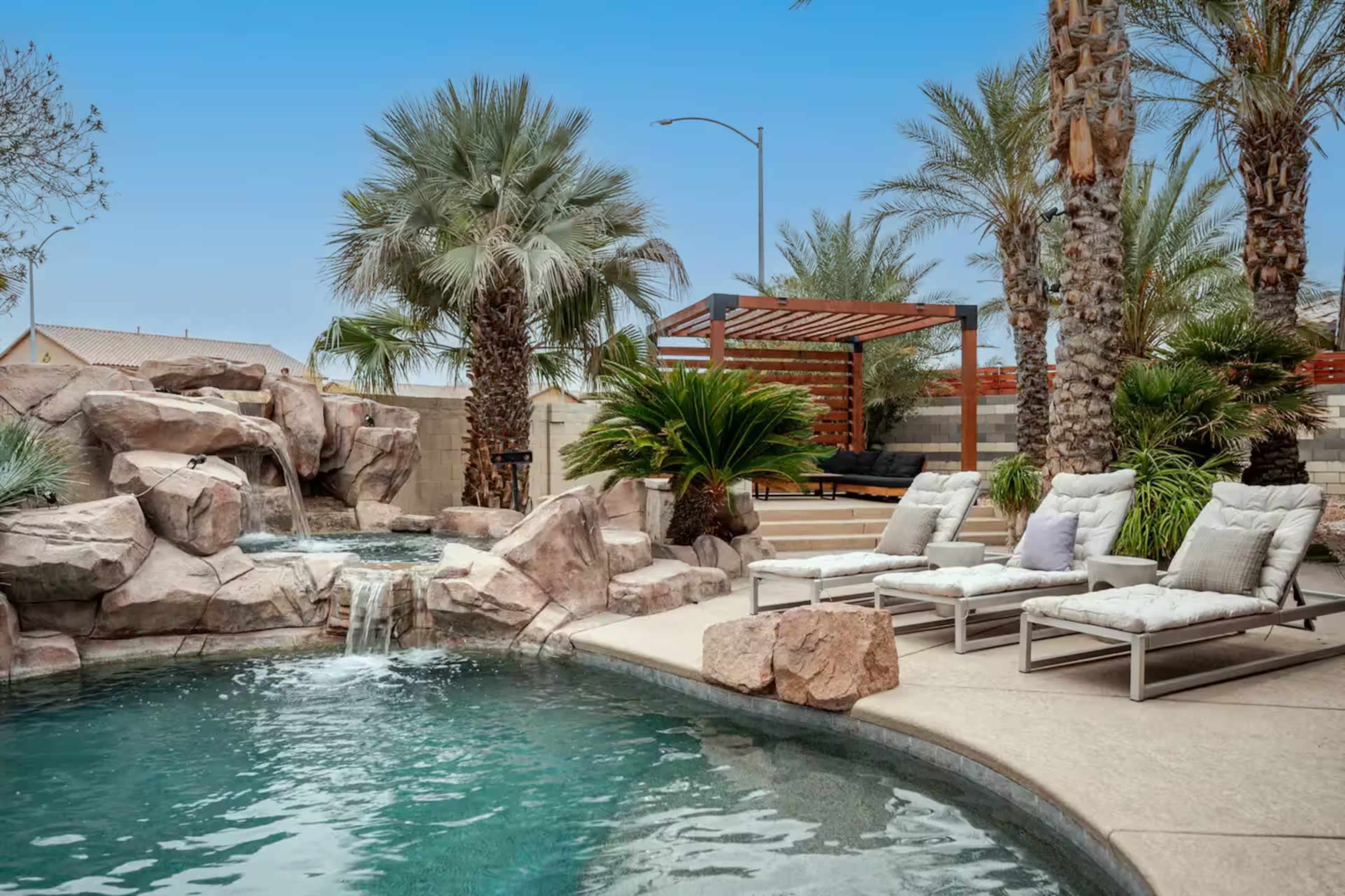 A backyard pool area features a rock waterfall, lounging chairs, and palm trees under a blue sky.