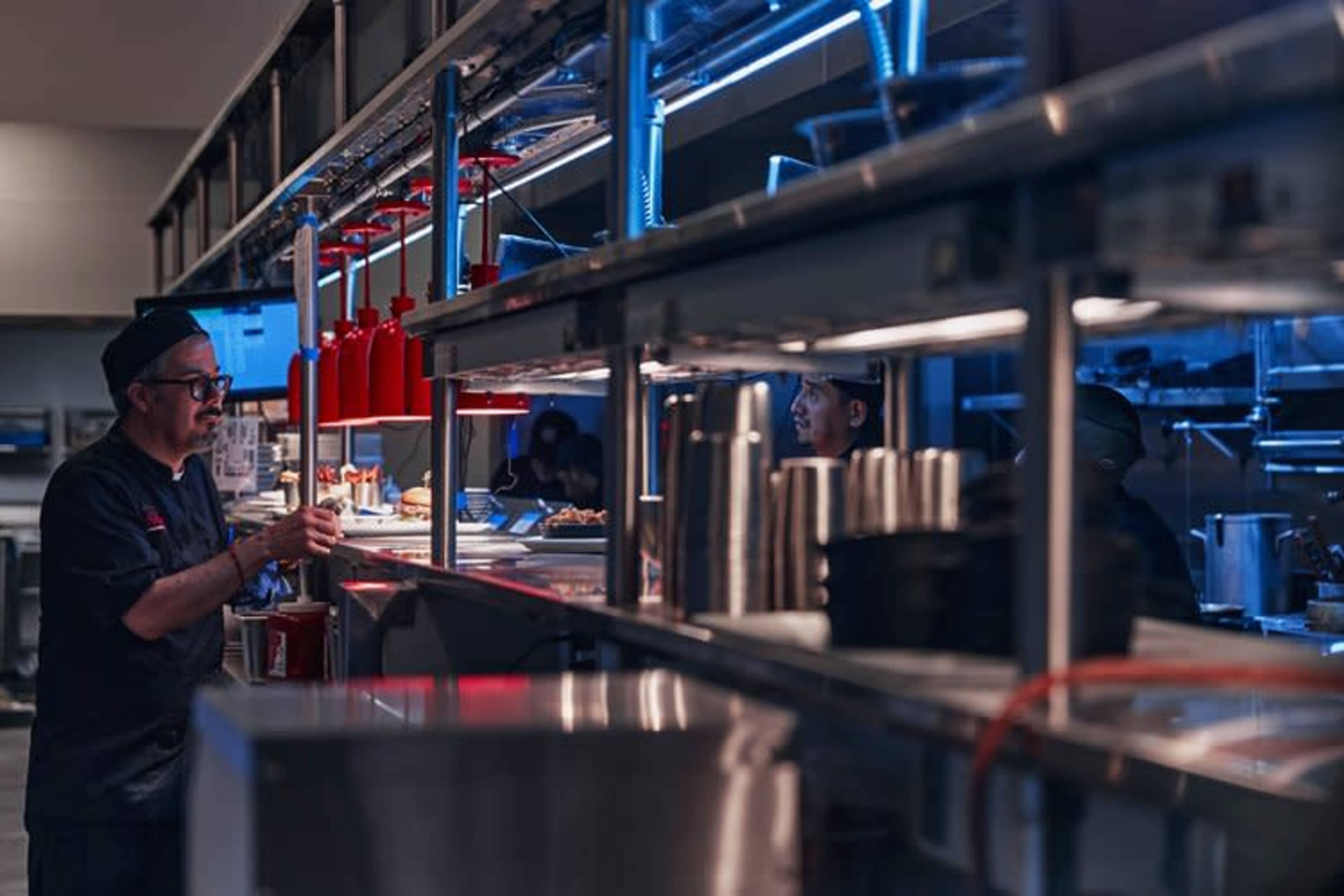 A chef stands behind a counter in a dimly lit kitchen, with illuminated red dispensers and various cooking equipment visible on the shelves.