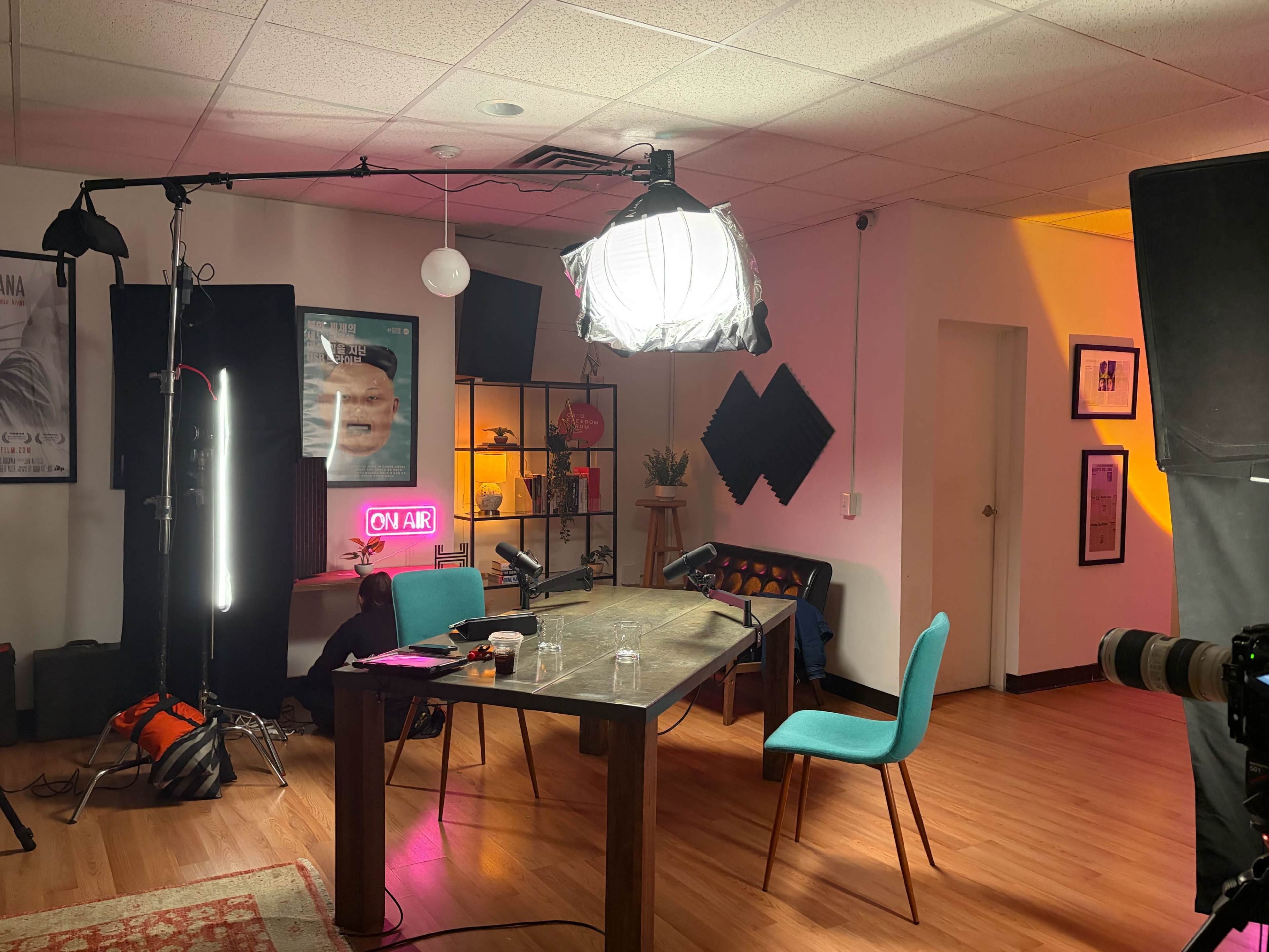 The image shows a well-lit podcast studio featuring a table with equipment, chairs, and various lighting setups against a backdrop of decorated walls.