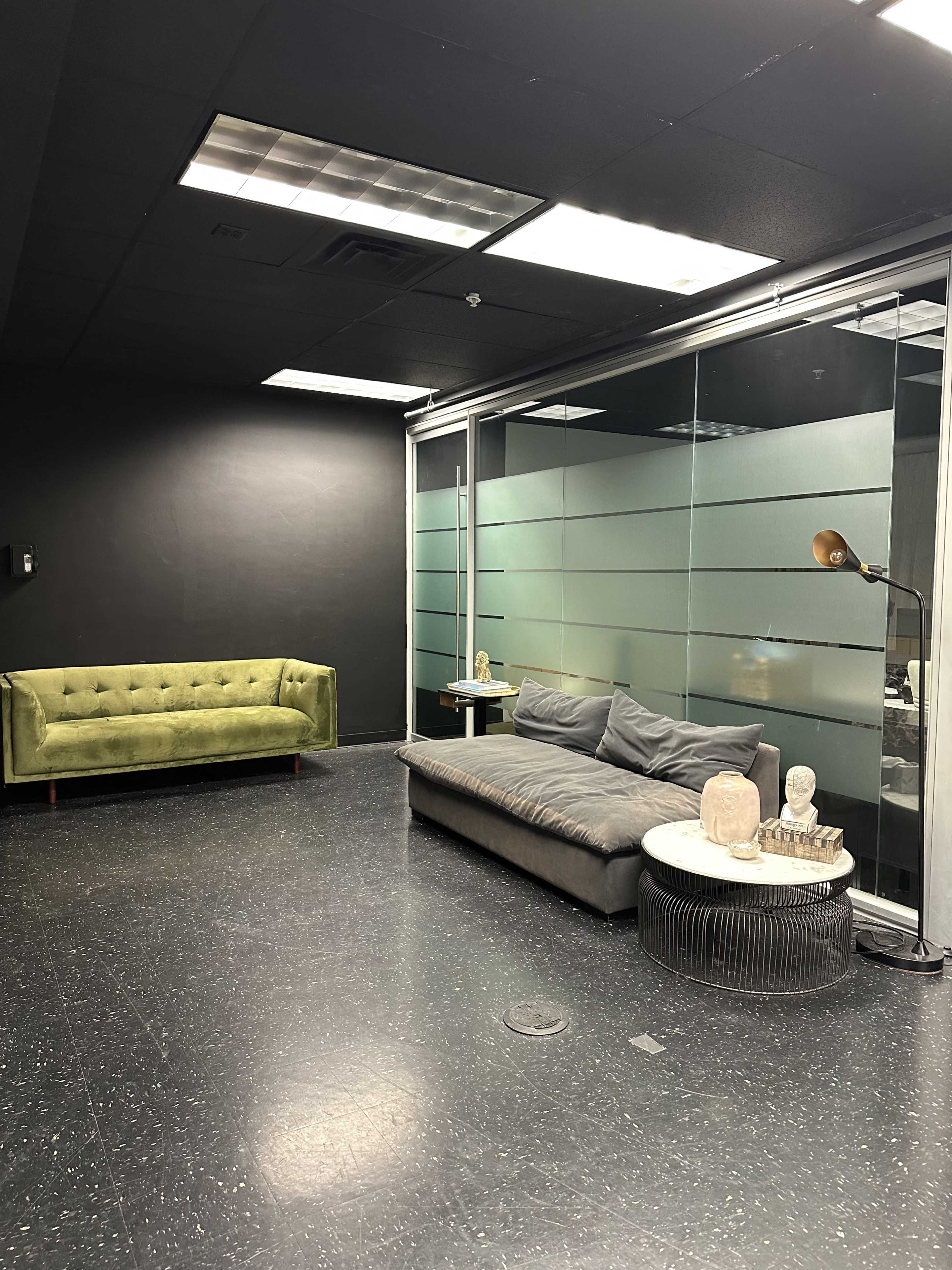 The image shows a modern office space with a green couch, a gray sofa, a round table, and glass partition walls.