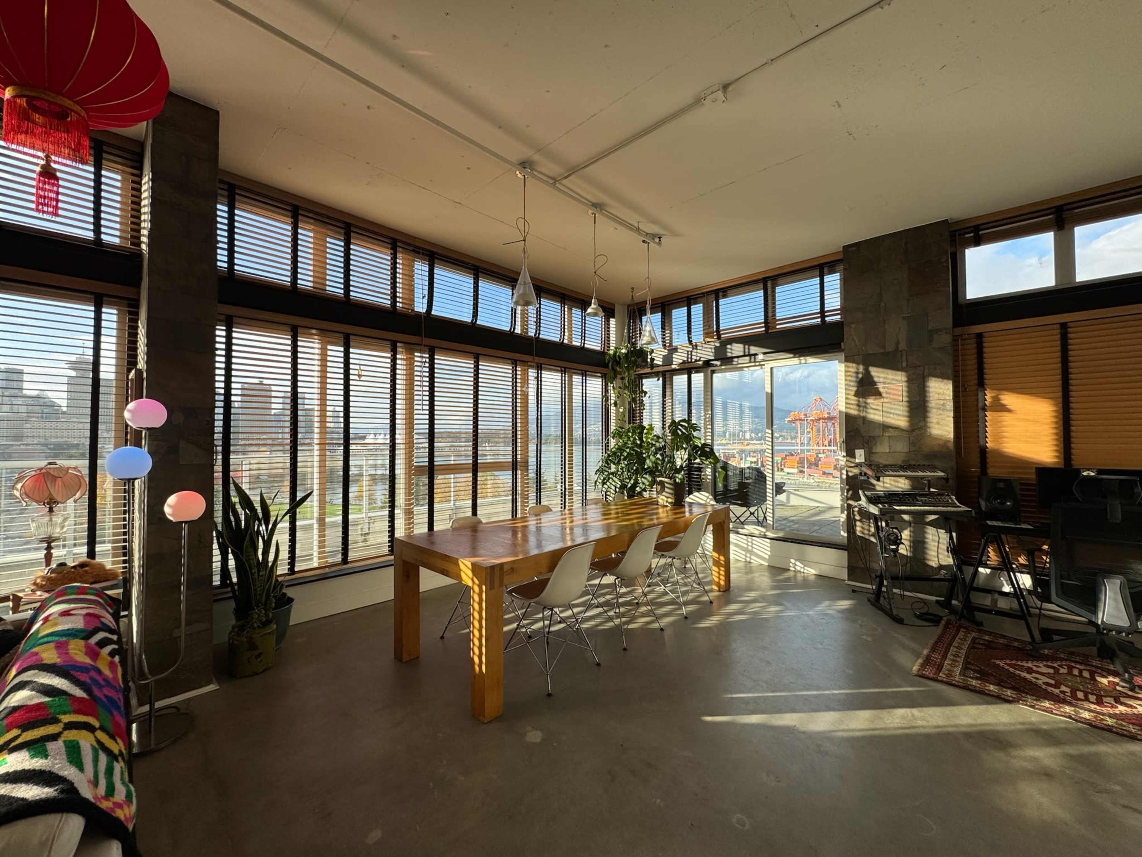 The image shows a modern dining area with a wooden table, white chairs, and large windows that offer a view of the city, featuring a mix of plants and decor.
