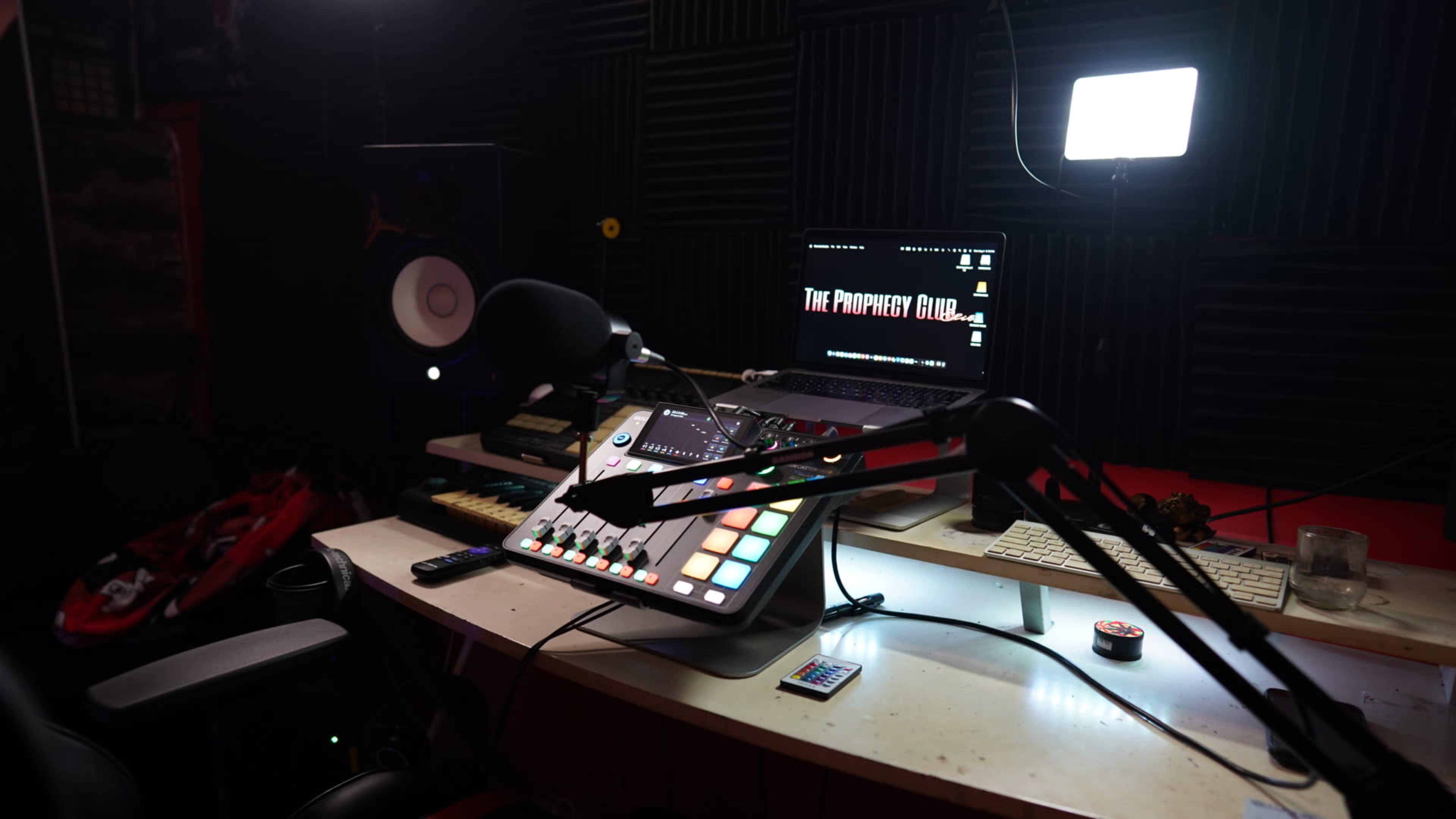 A music production workspace features a mixing console, a computer monitor displaying "The Prophecy Club," and studio speakers amidst a dark background.