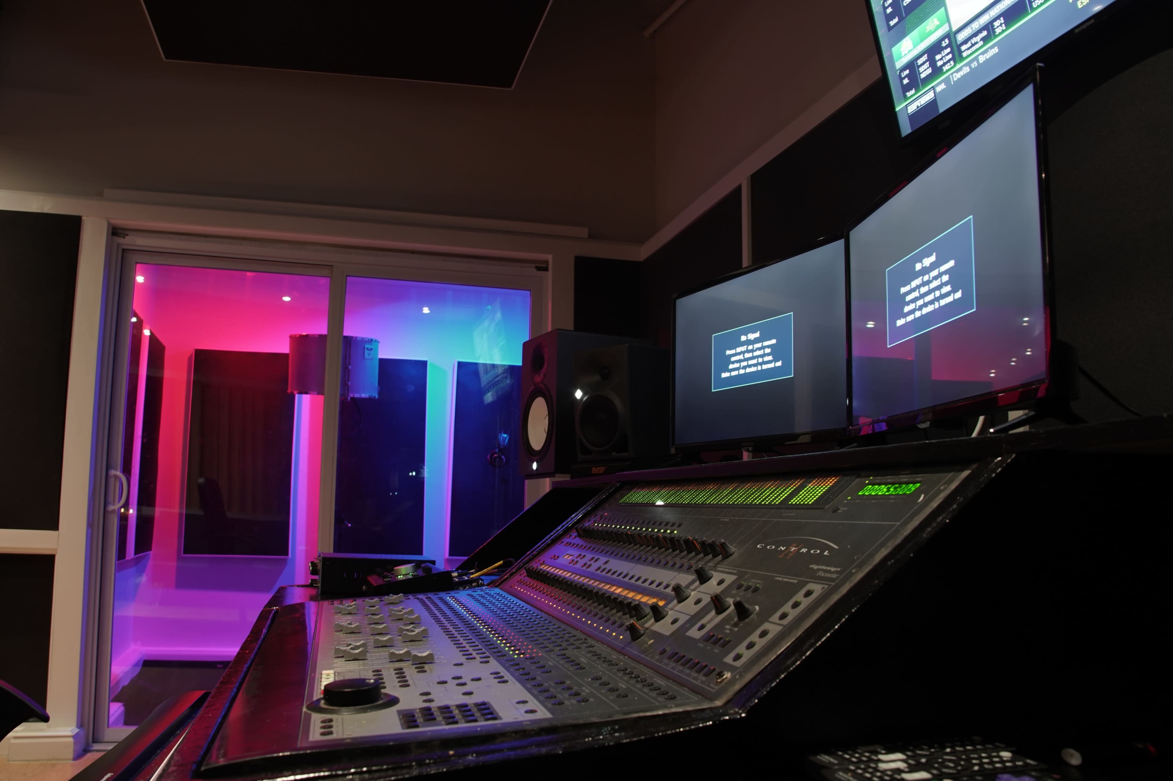 A music recording studio with a control desk, multiple screens, and a glass-enclosed vocal booth illuminated with colored lights.