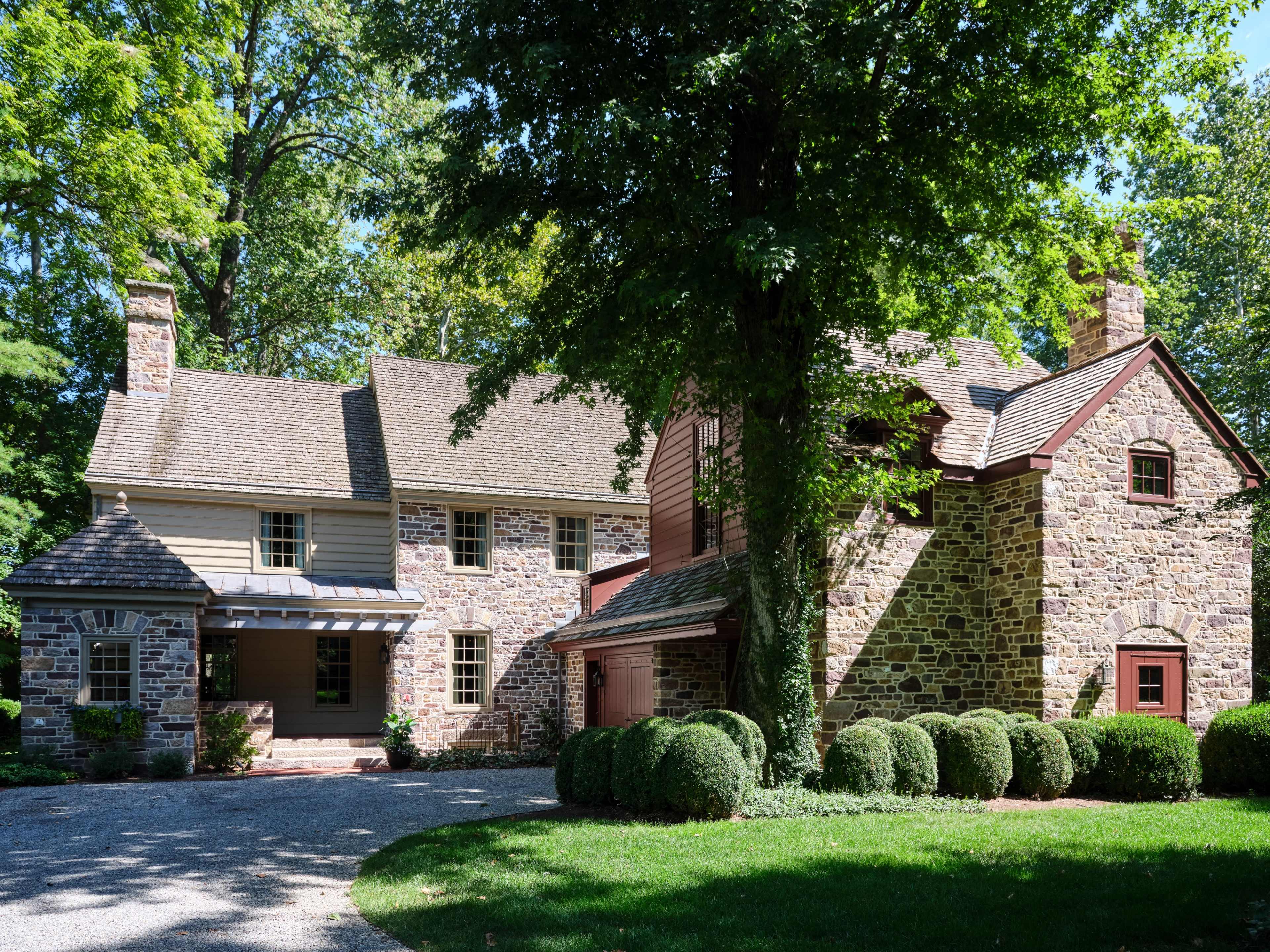 A stone and wood house with a gravel driveway, surrounded by lush greenery and trimmed hedges.