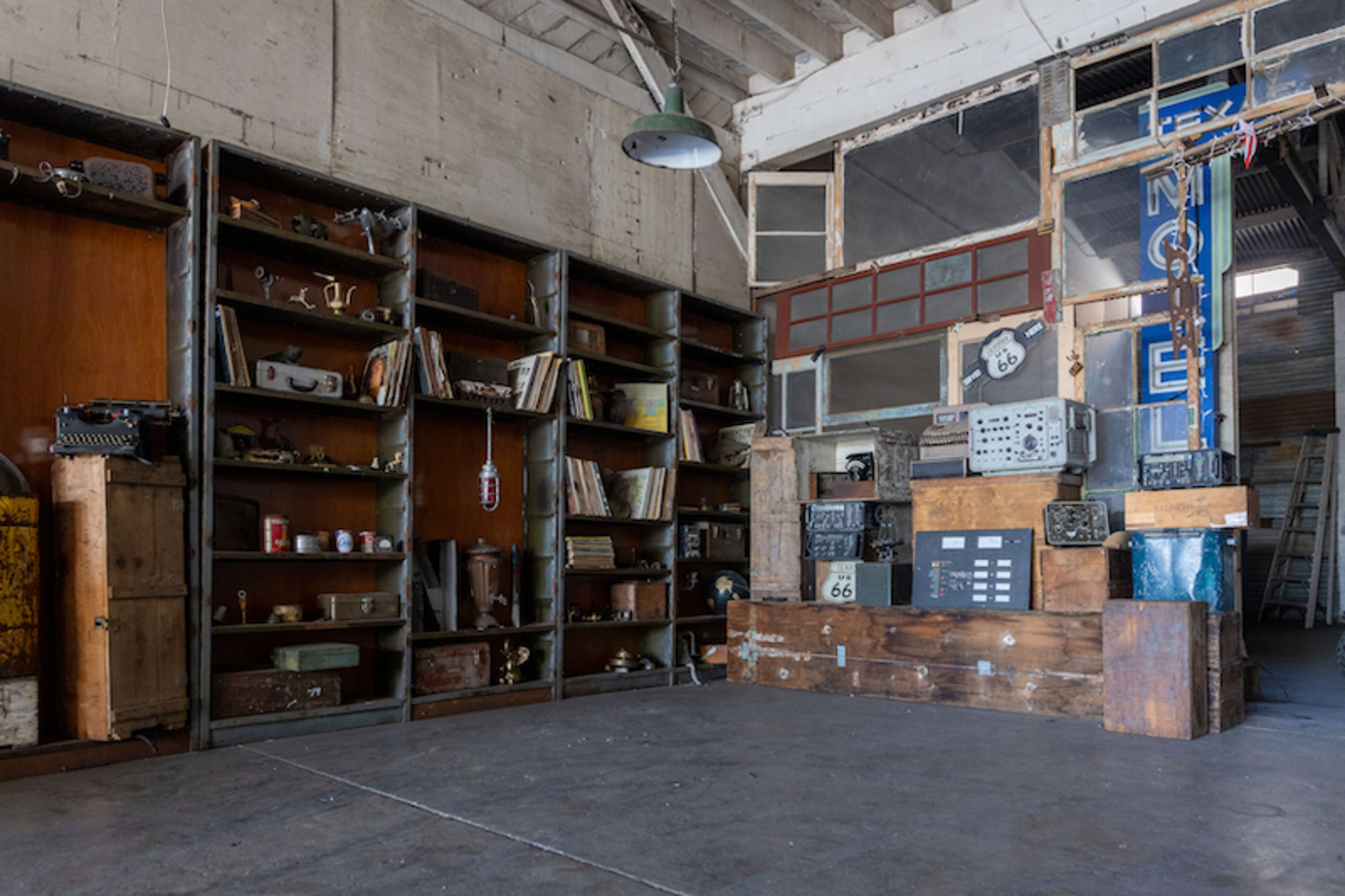 A spacious room featuring wooden shelves filled with books and various items, alongside a large wooden display structure against an industrial backdrop.