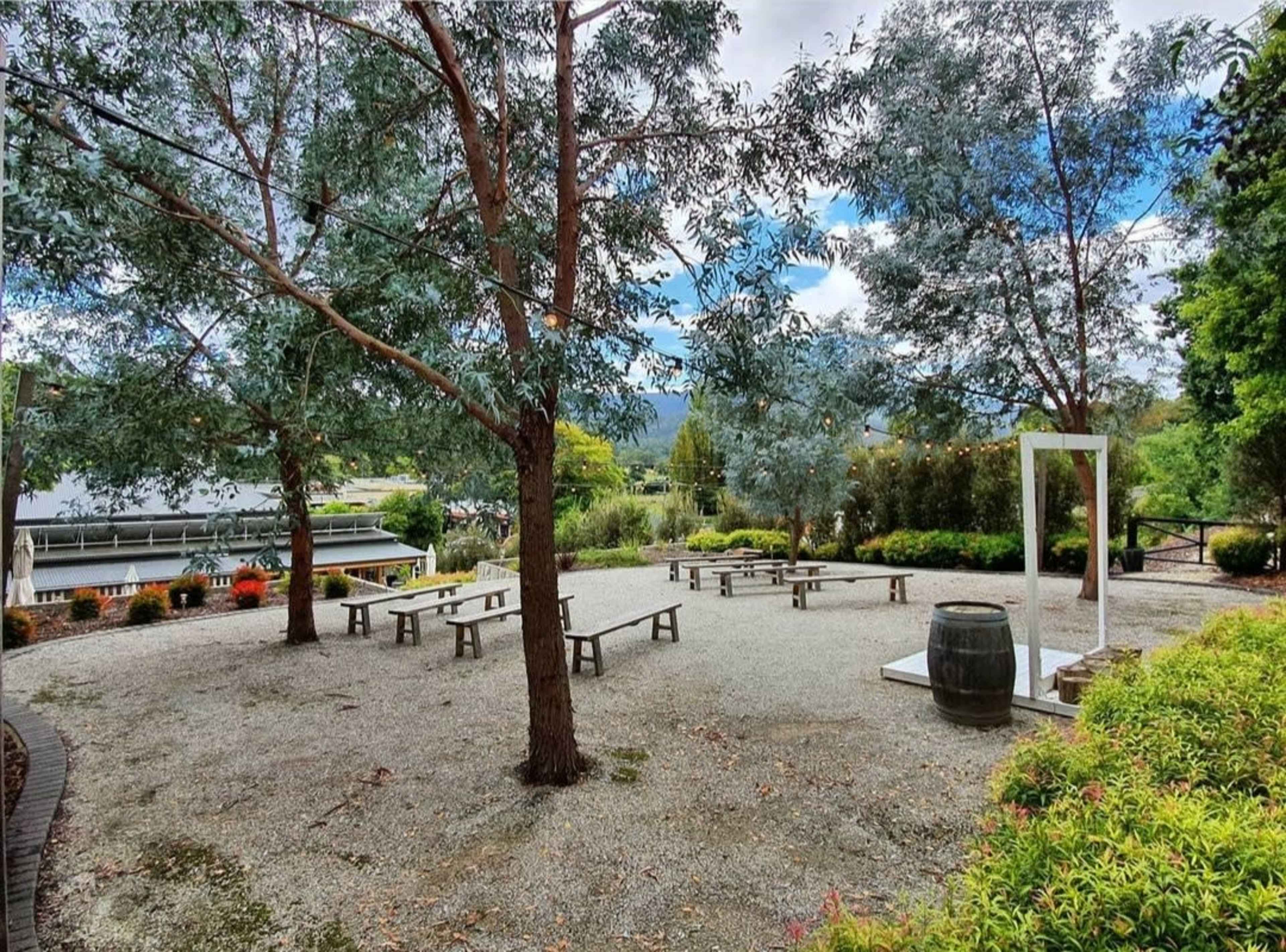 A landscaped outdoor area with gravel ground, wooden benches arranged around a central space, and several trees providing shade.