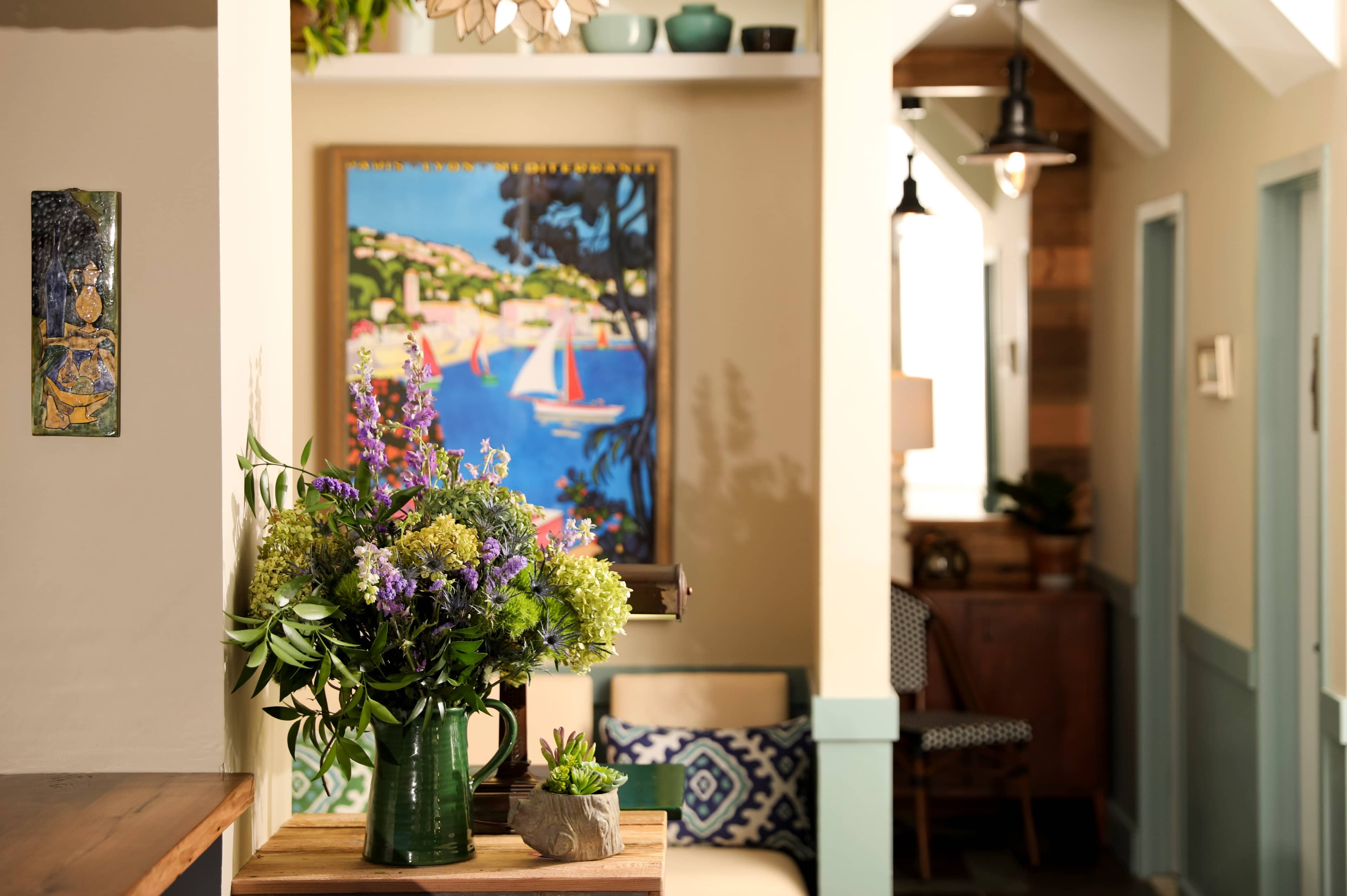 A brightly lit interior hallway featuring a vase of flowers on a wooden table, with a colorful painting and various decorative items on the walls.